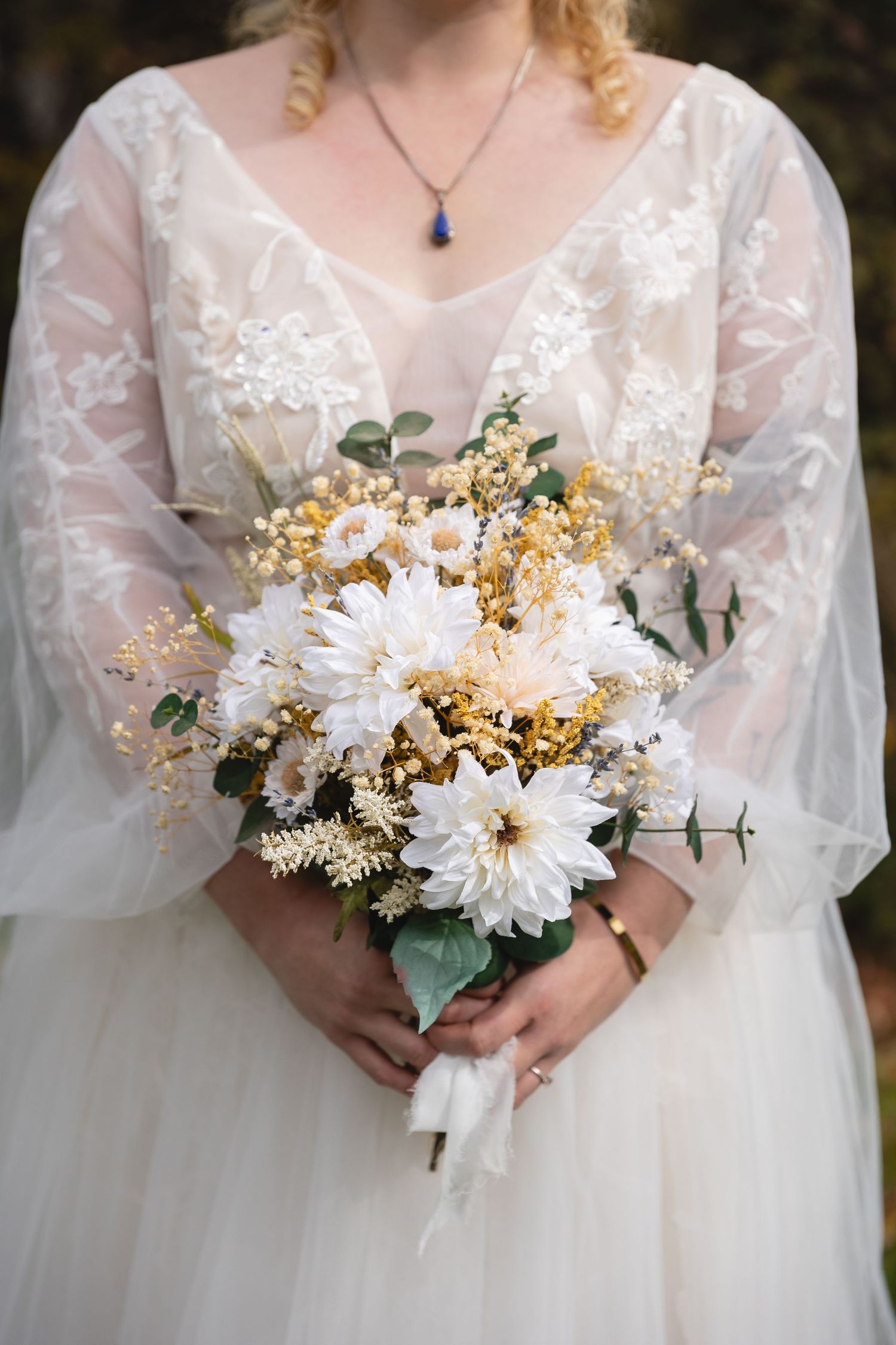 A bridal bouquet of white dahlias and yellow flowers with delicate lace dress details and blue pendant necklace.