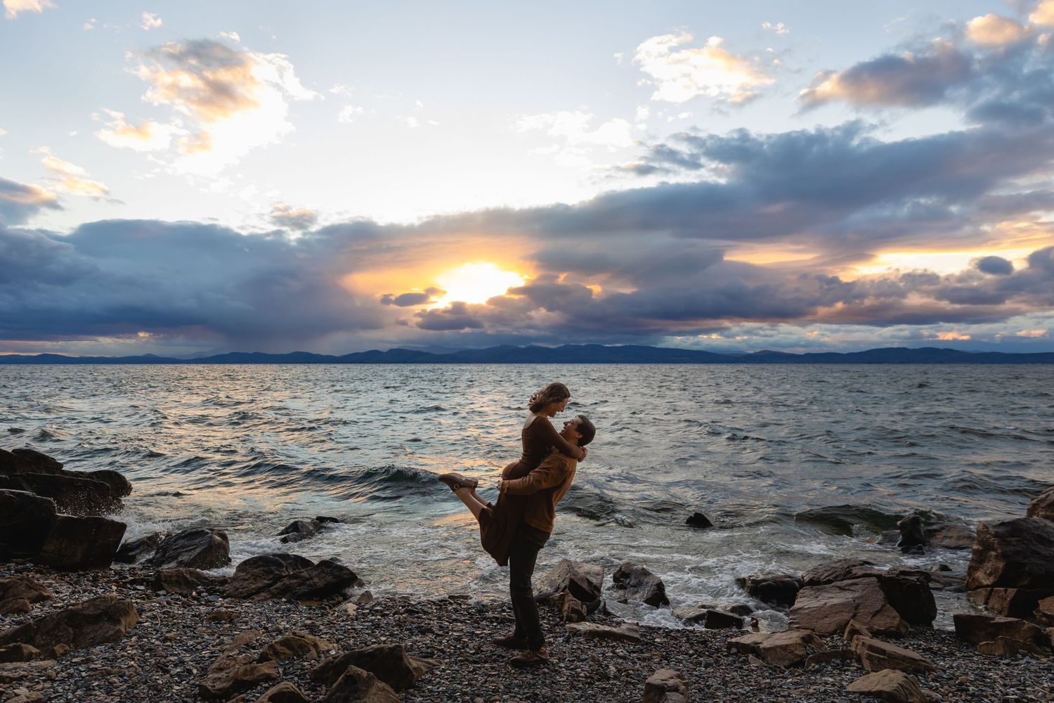 Silhouetted couple embracing on rocky beach during dramatic sunset over ocean with cloudy sky.
