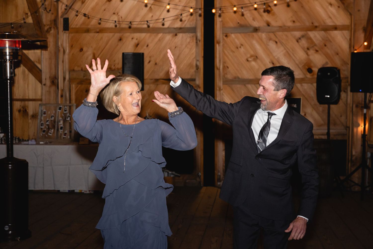 Two people dance together with raised arms in a rustic wooden barn reception venue with string lights on the ceiling.