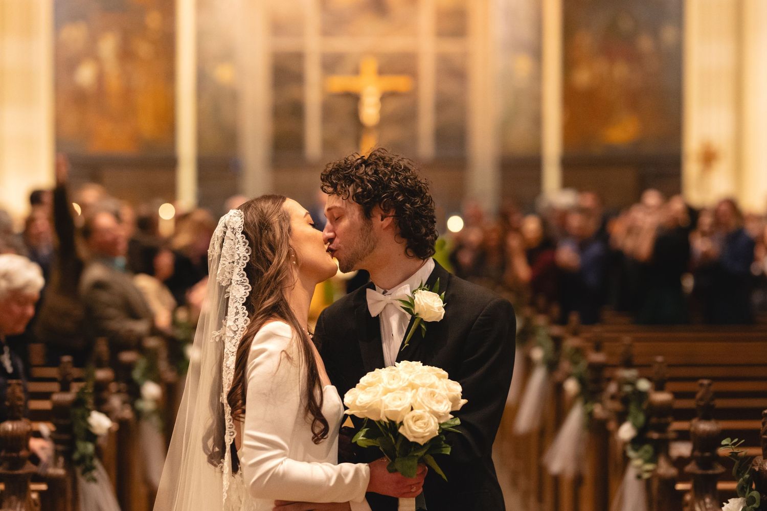 A romantic moment between newlyweds sharing a kiss at the altar while holding a white bouquet in a grand church setting.