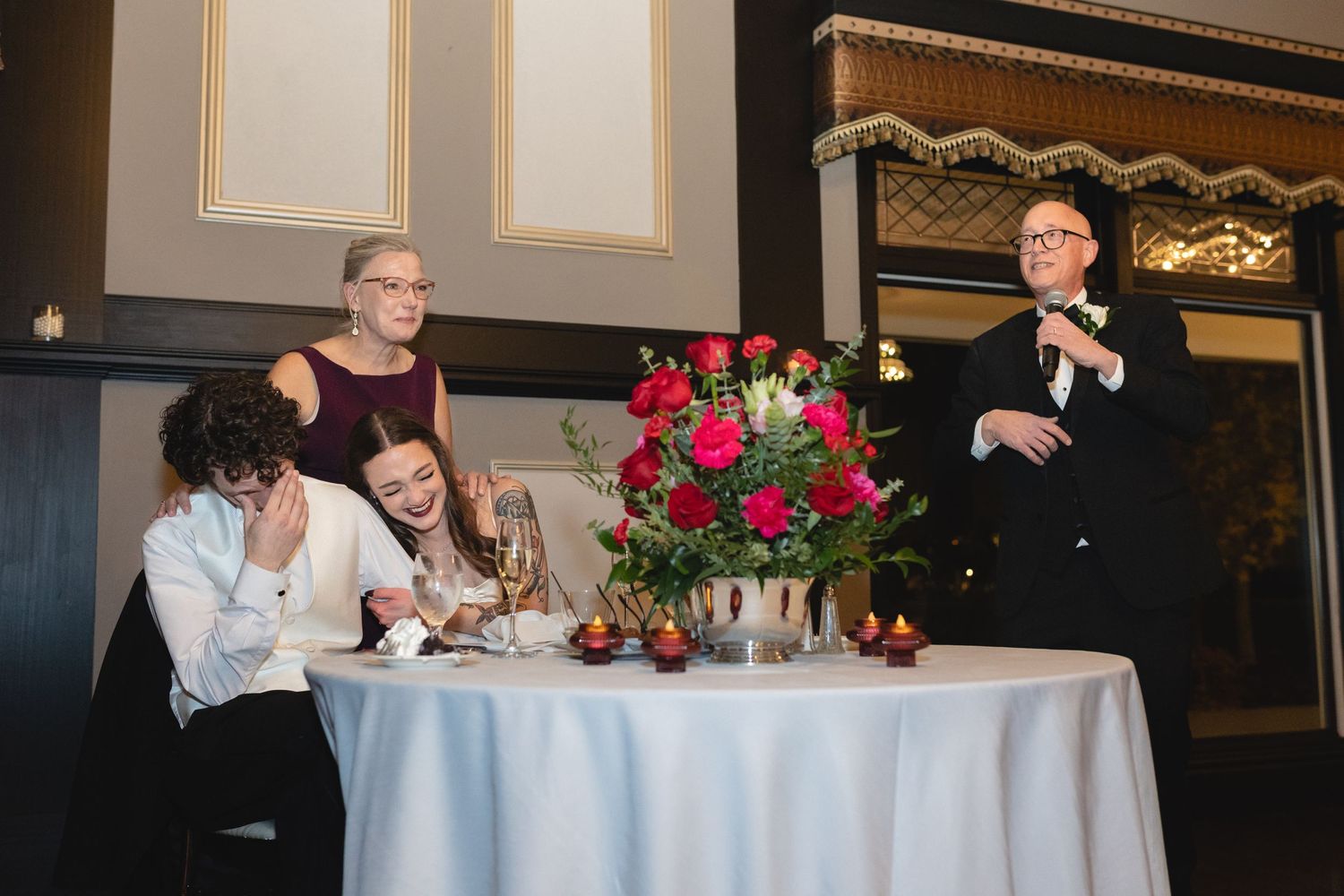 Guests gather around an elegant round table with a bright red floral centerpiece at a formal dining event.