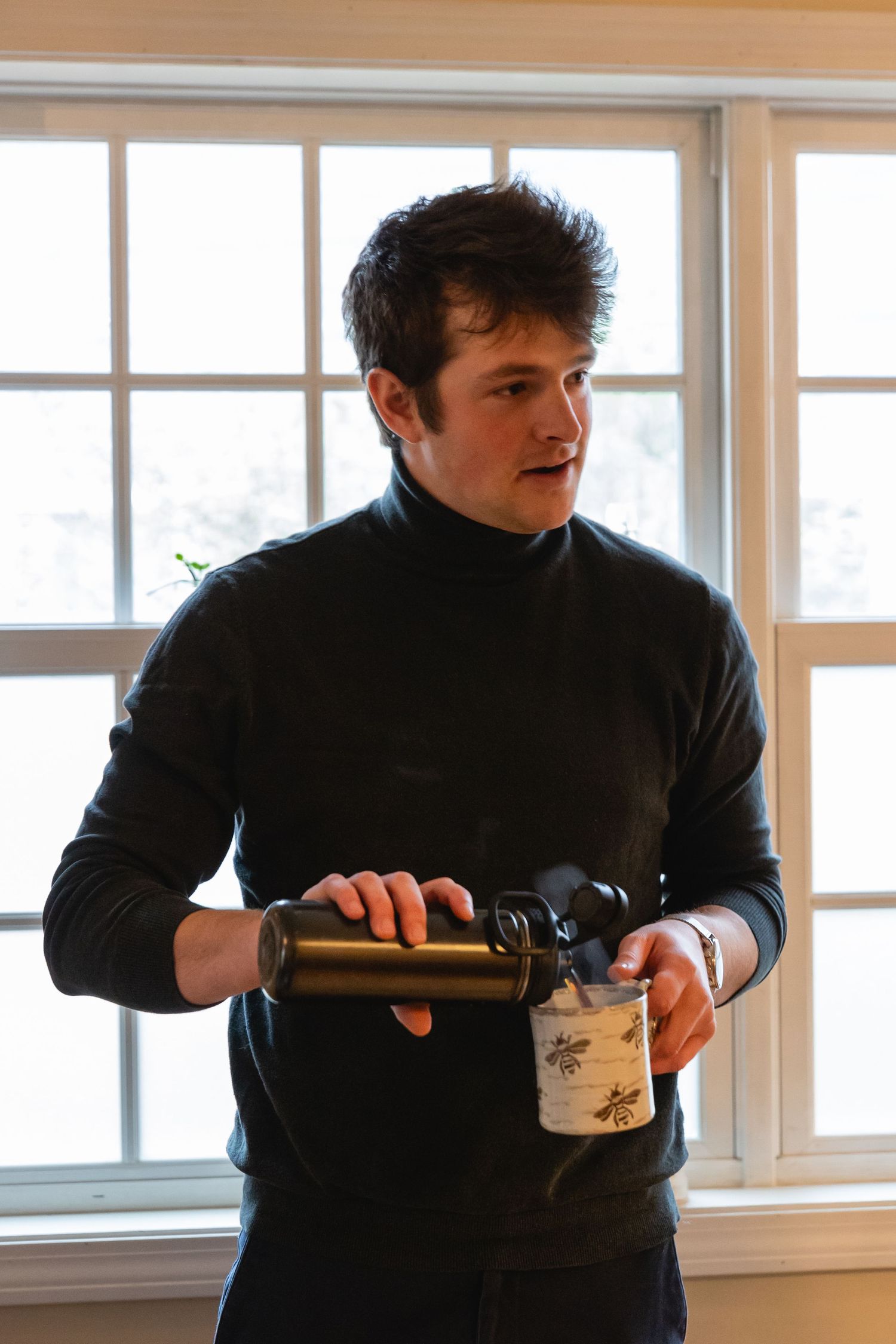 A person wearing a black turtleneck sweater pours a drink by a bright window.