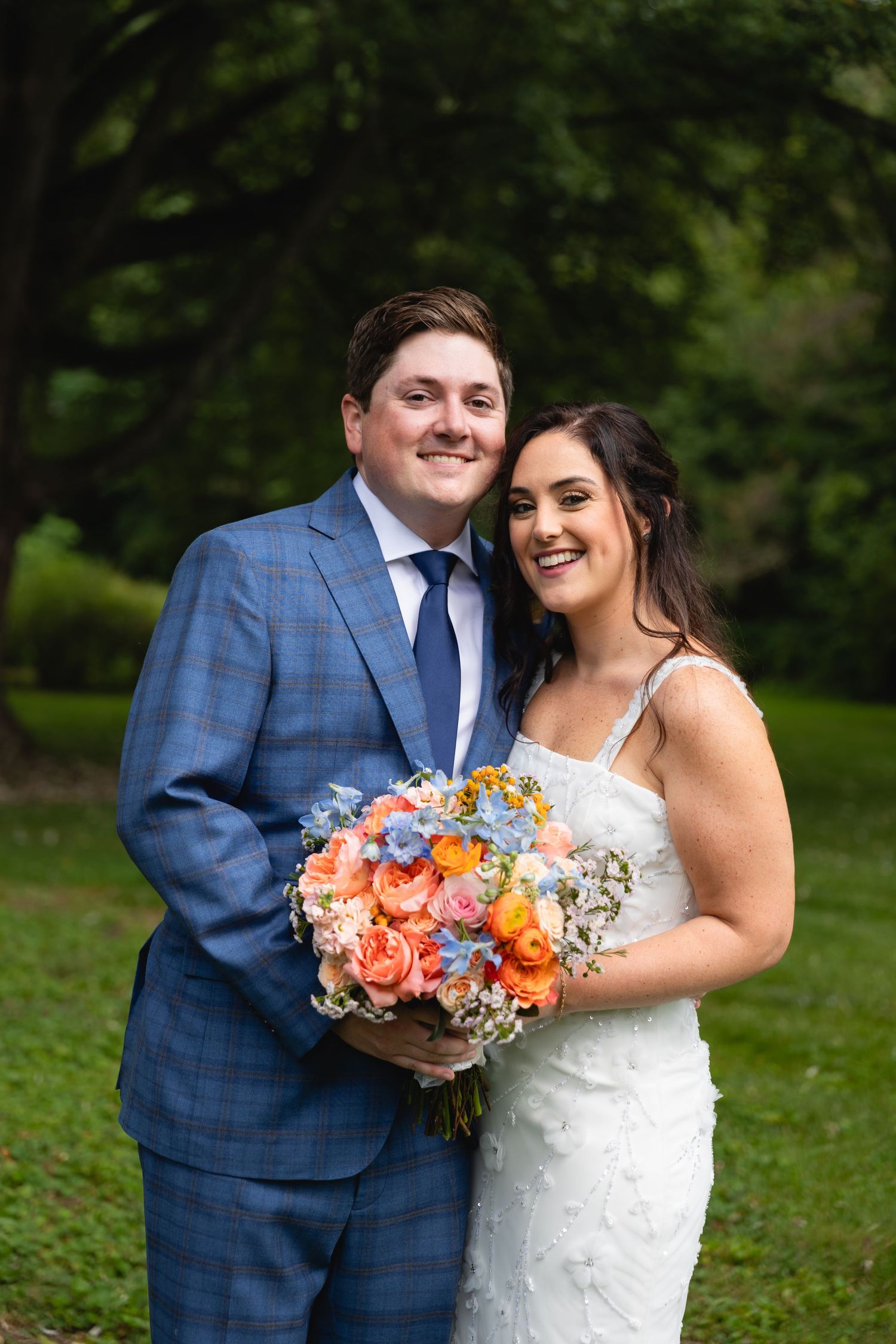 Newlywed couple poses outdoors with vibrant coral and orange bouquet in front of green foliage.