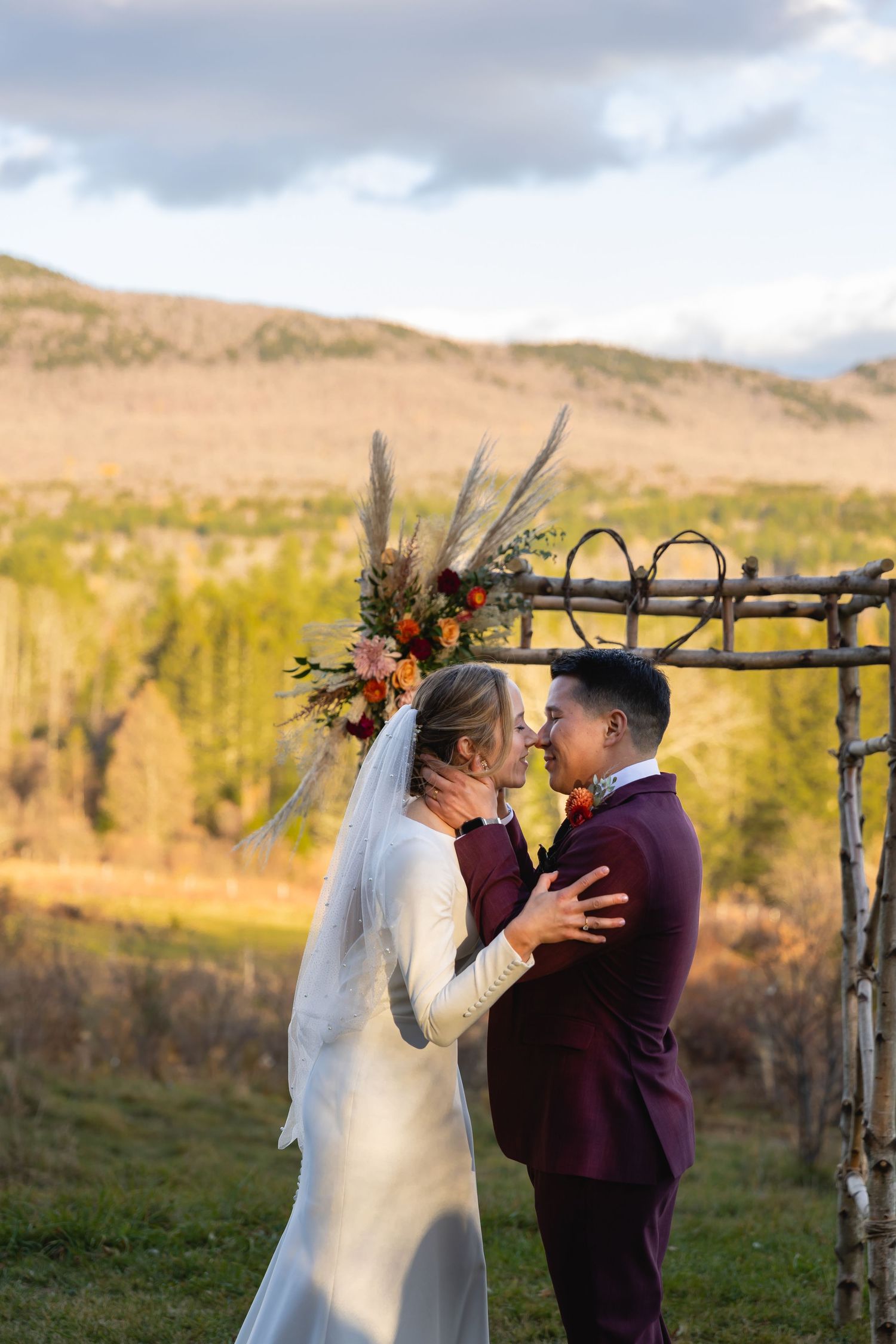 A mountainous outdoor wedding ceremony featuring a rustic wooden arch decorated with dried flowers and autumn florals.