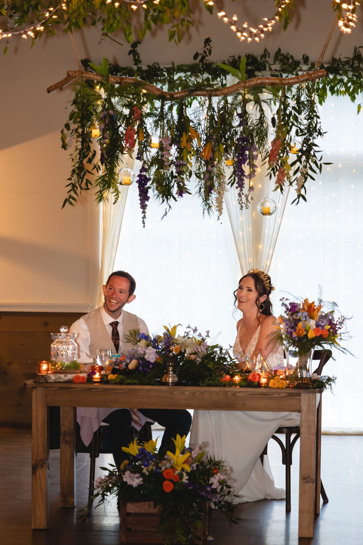 Wedding couple seated at rustic head table with hanging floral installation and twinkle lights above.