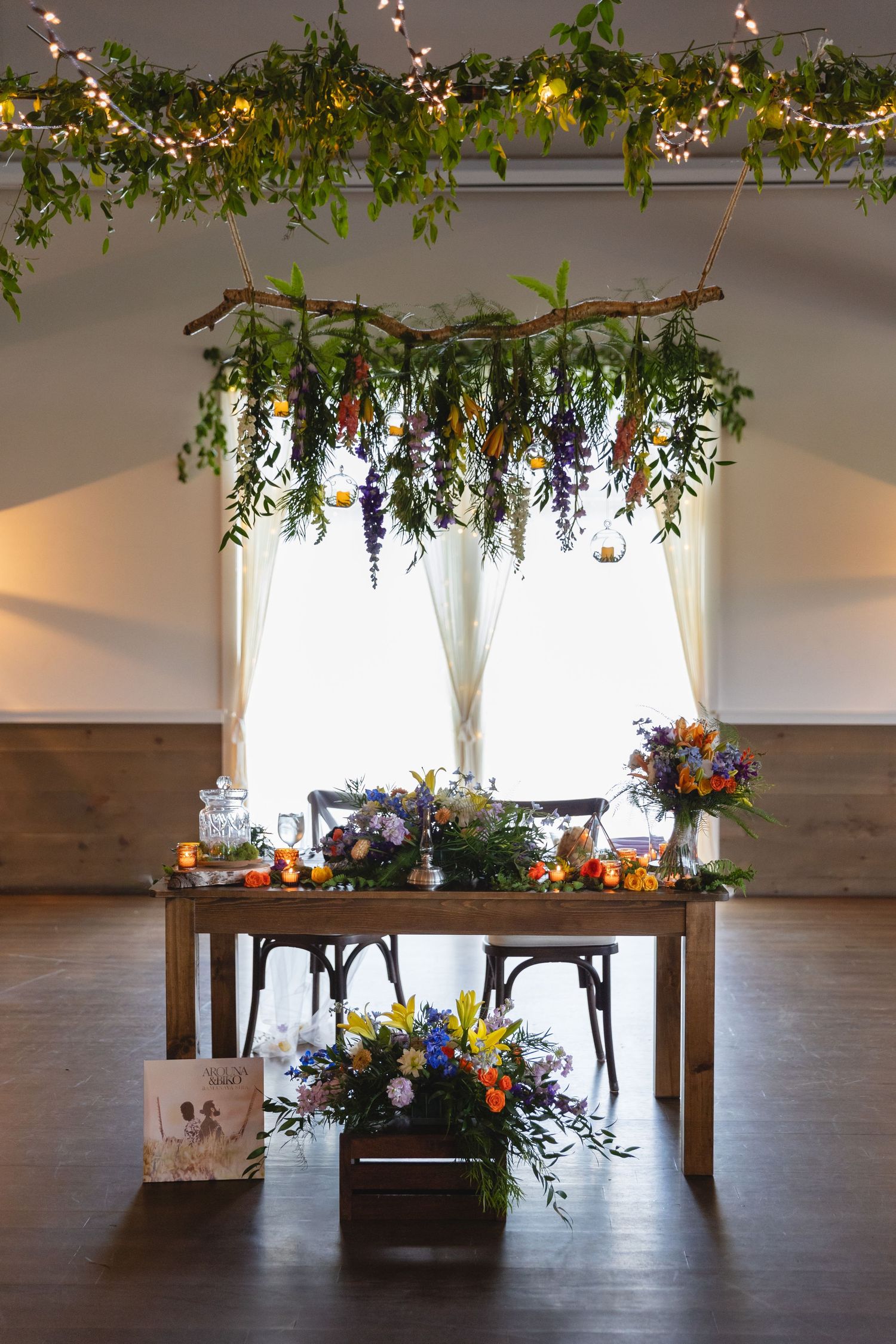 Wedding table setup with hanging floral installation, string lights and rustic wooden decor.