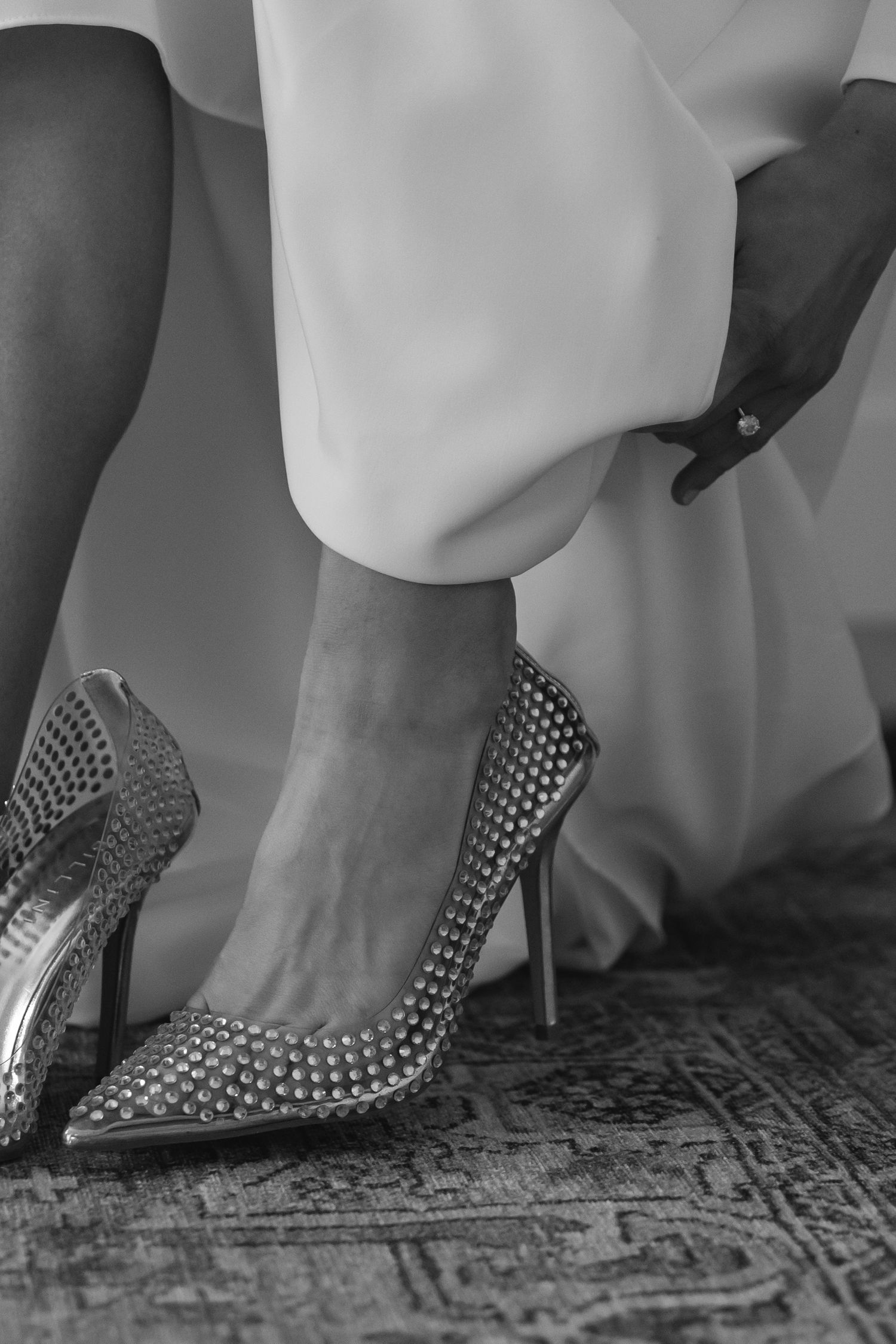 Close-up black and white photo of feet wearing studded silver high heel pumps on a patterned carpet.