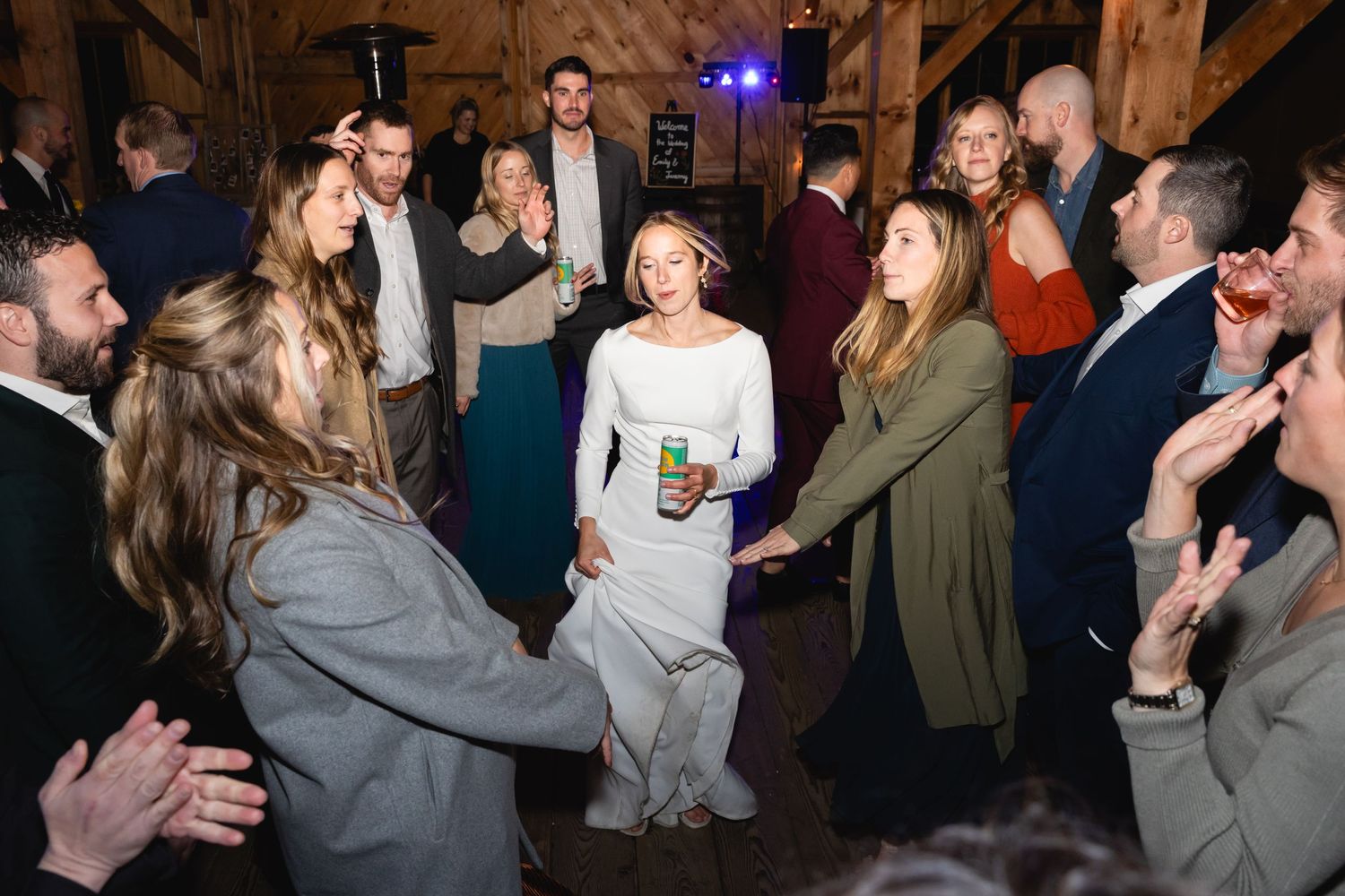 Wedding guests form a circle on the dance floor around a smiling dancer in white during a lively reception celebration.