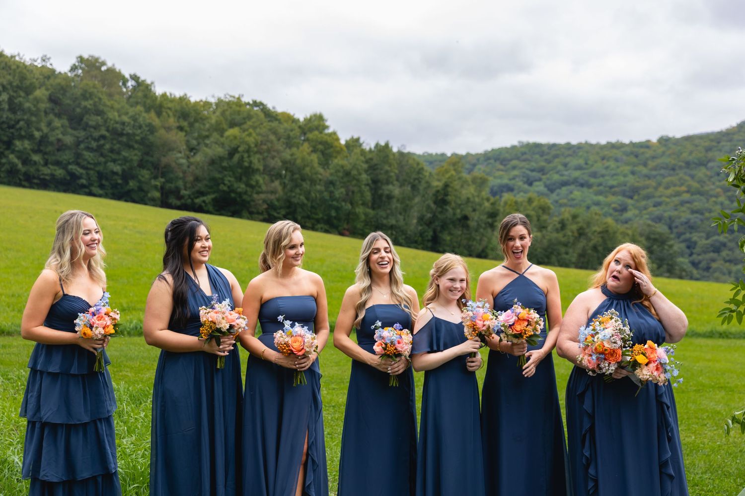 A group in long navy blue bridesmaid dresses holding colorful bouquets pose together on a lush hillside.