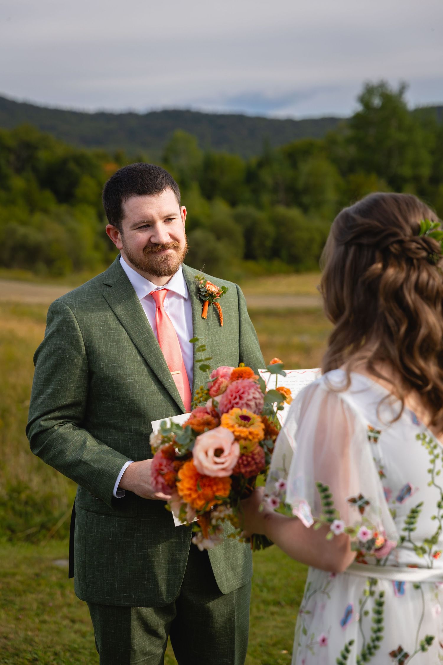 A couple exchanges vows in a field surrounded by wildflowers and mountains during an outdoor ceremony.