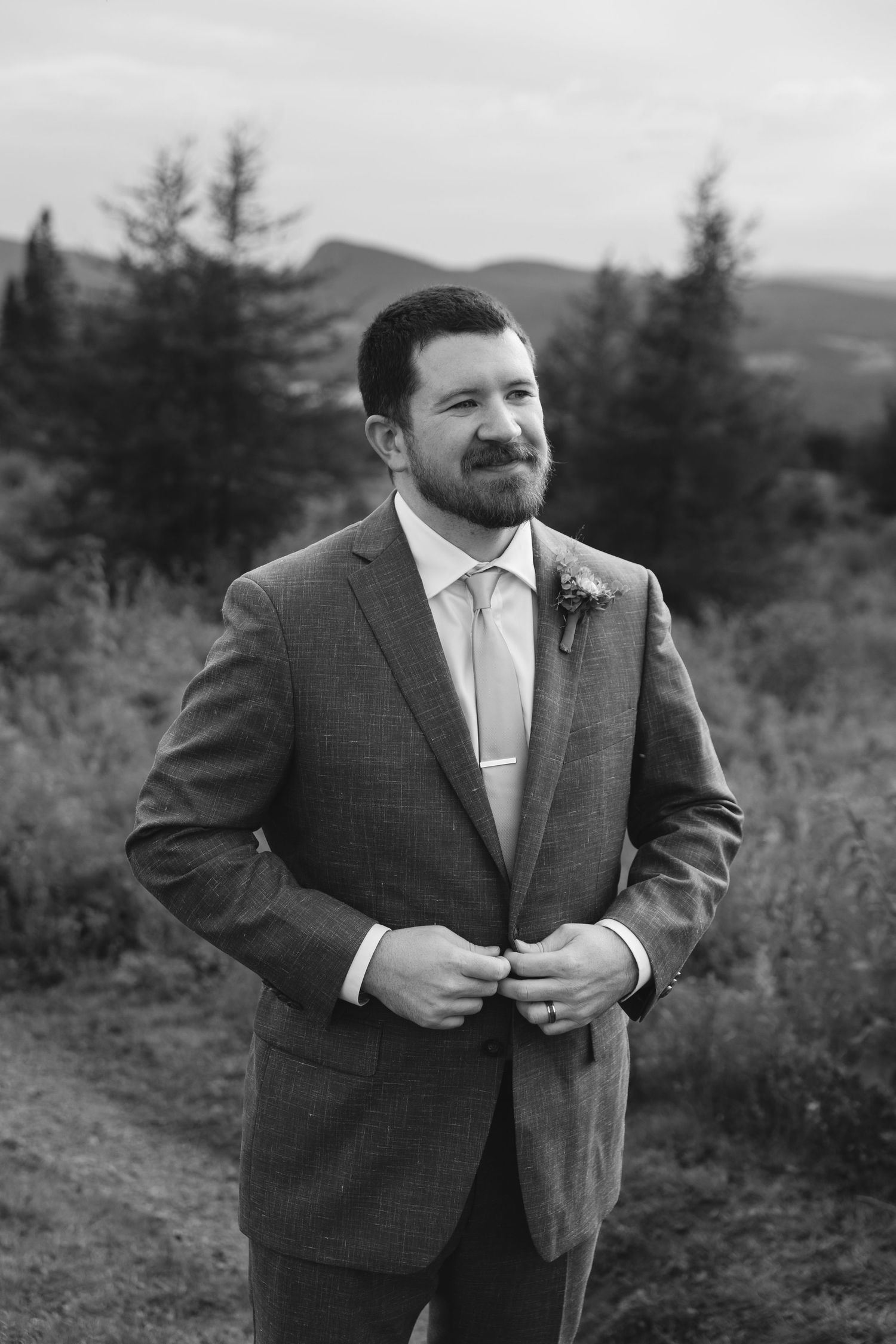 A person in a dark suit jacket stands outdoors against a scenic mountain landscape background in black and white.