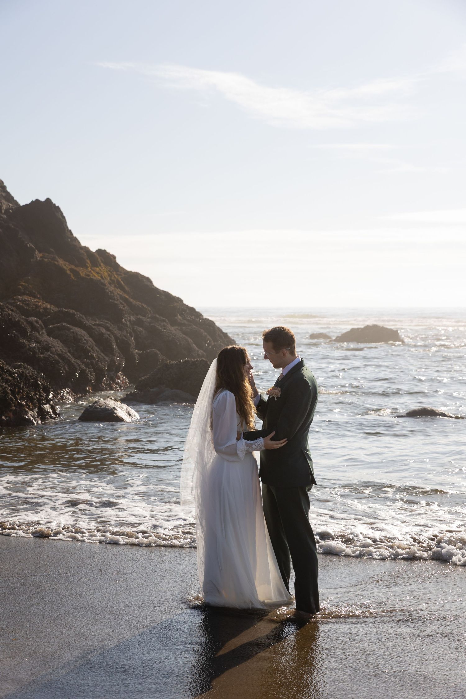 A couple in wedding attire stands together on a rocky beach at sunset with waves crashing behind them.