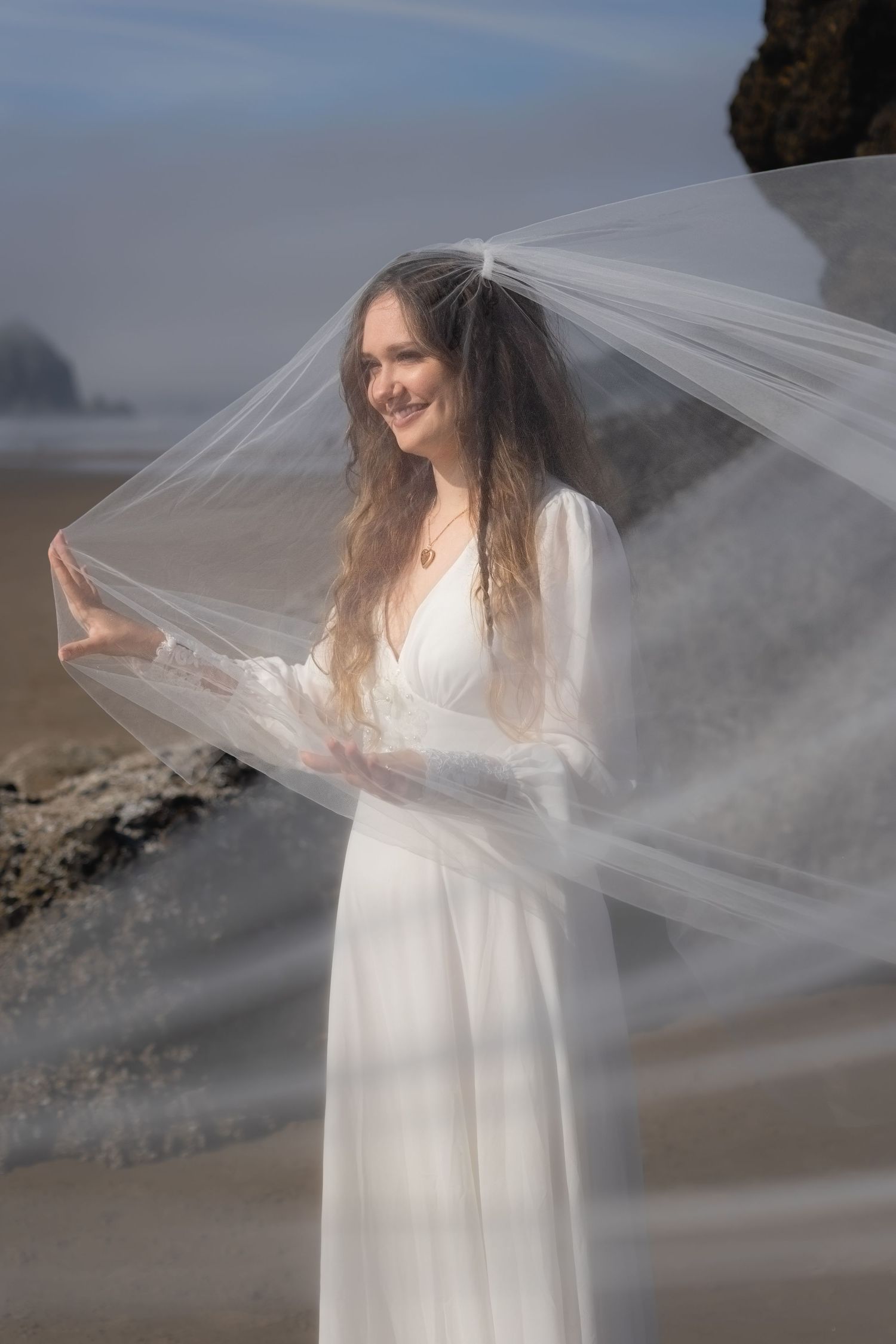 A bride in a flowing white dress stands on a misty beach with her veil floating in the breeze.