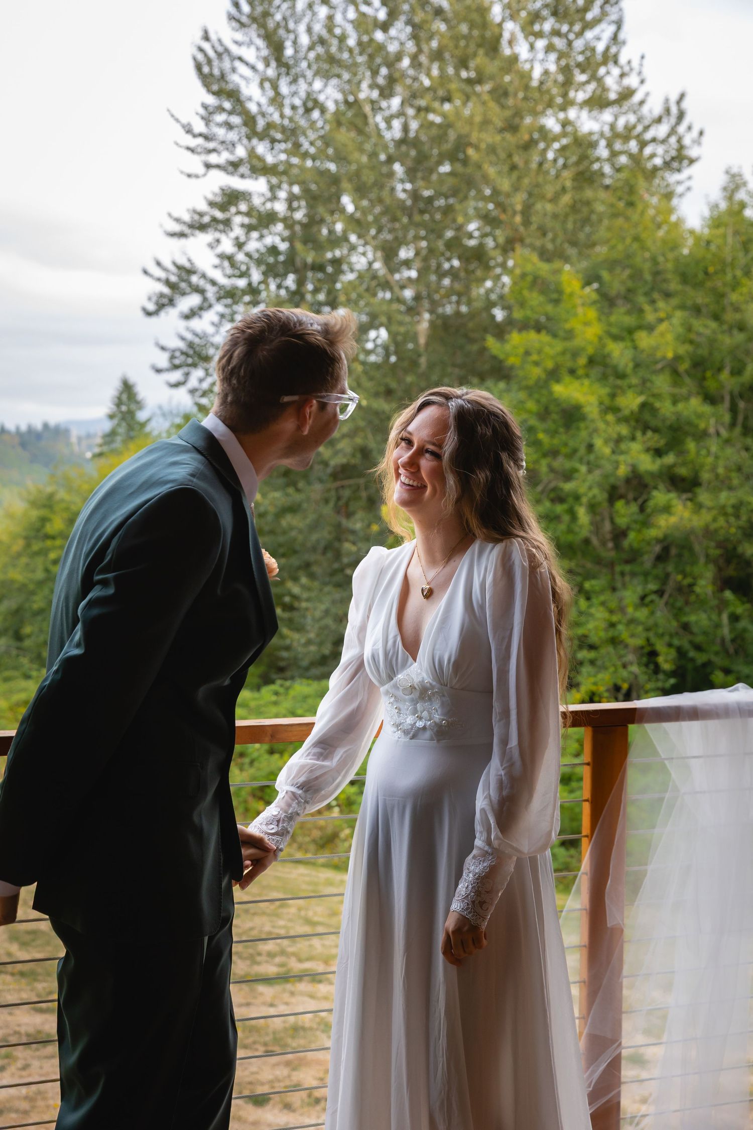 A couple in wedding attire share a tender moment on a deck overlooking a scenic forest landscape.