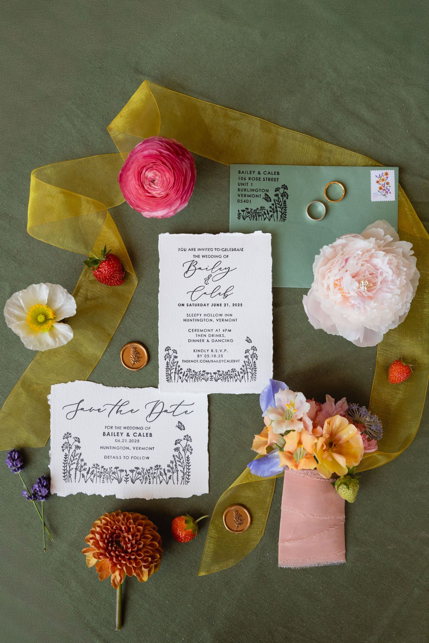 Wedding invitation suite with delicate floral details and calligraphy laid out on a dark green background with scattered flowers.