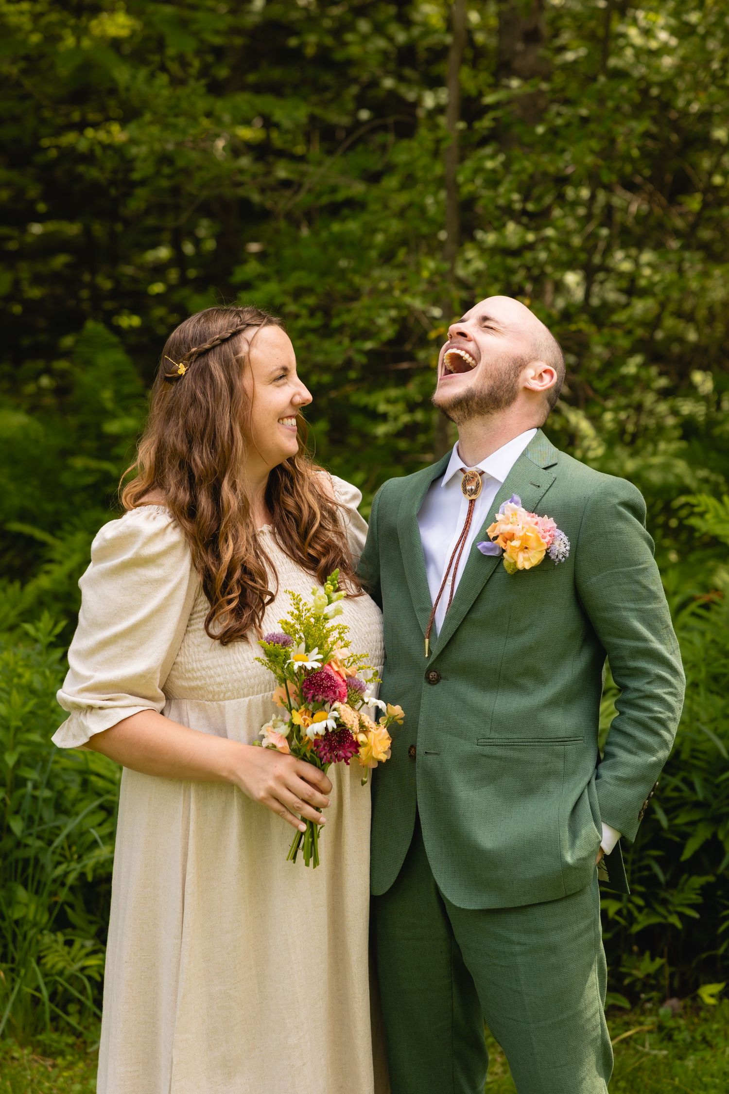 A couple in wedding attire sharing a joyful laugh together in a lush green outdoor setting.