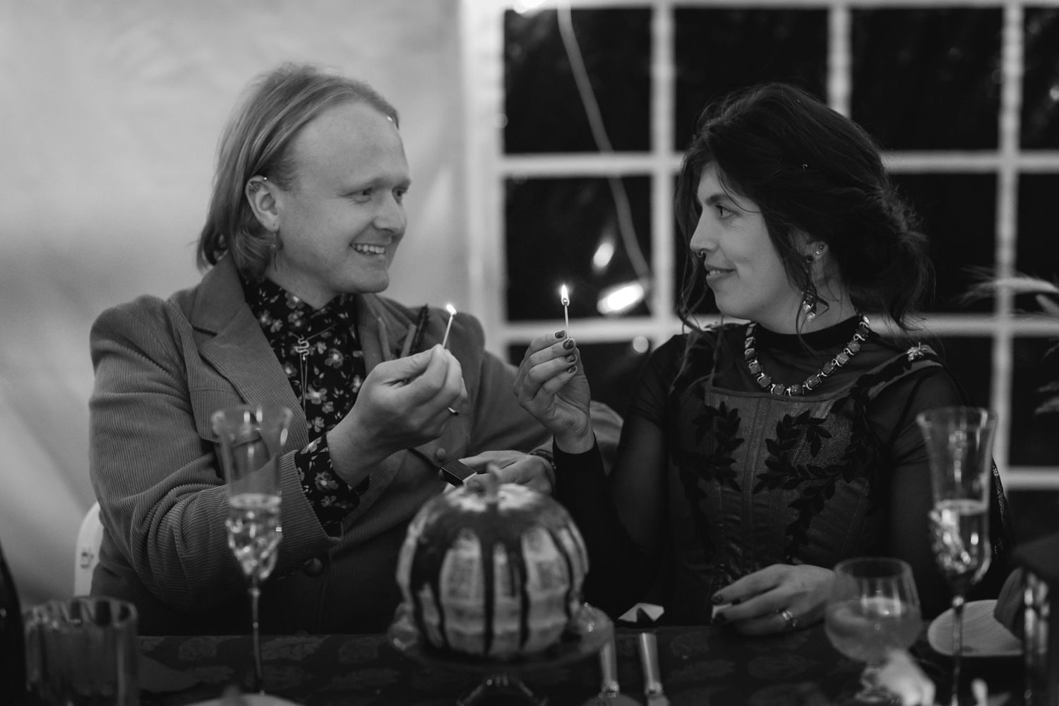 A black and white photo of two people having an animated conversation at a table with wine glasses and a decorative vase.