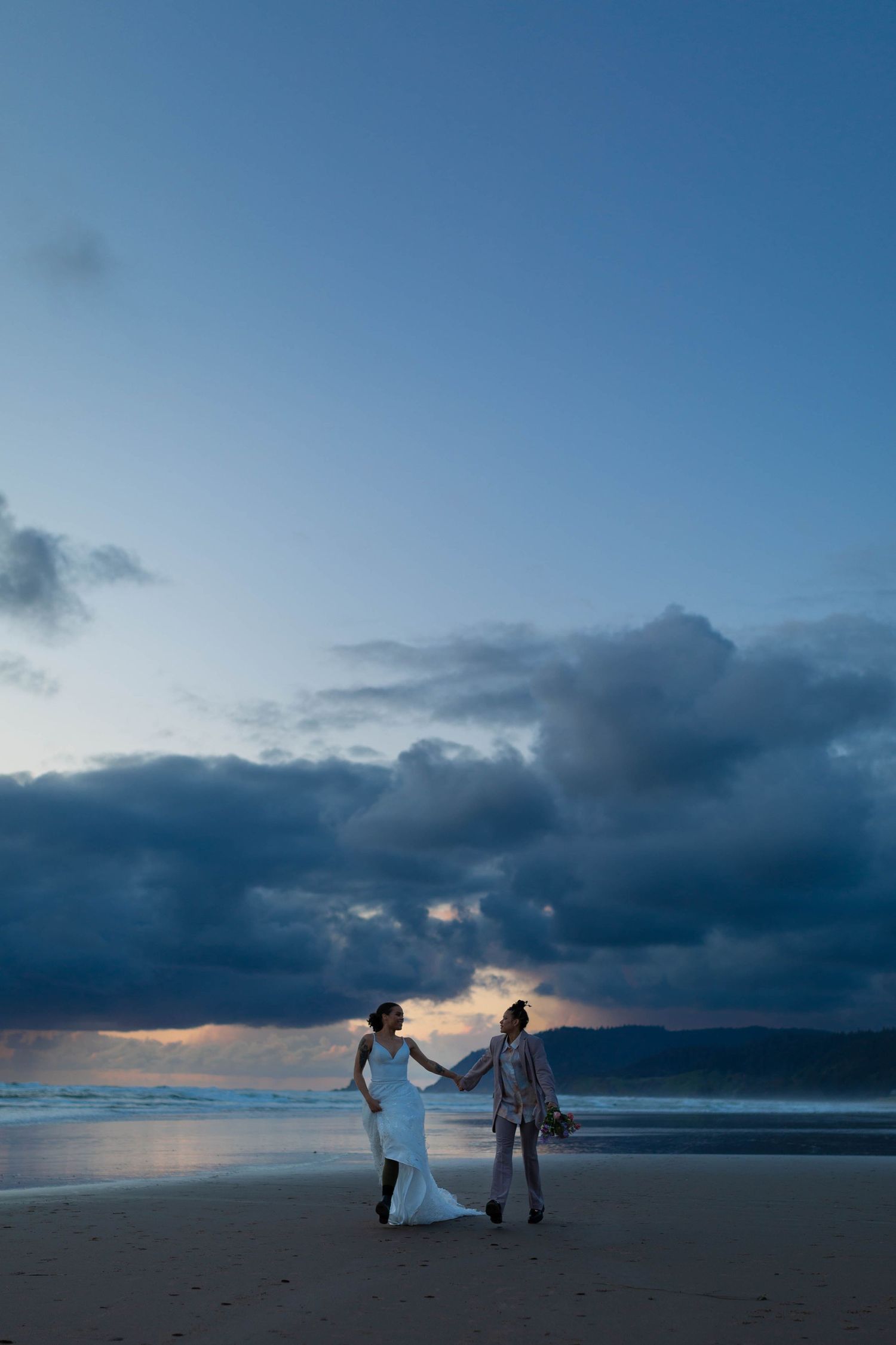 A couple walks hand in hand on a beach at sunset with dramatic storm clouds overhead.