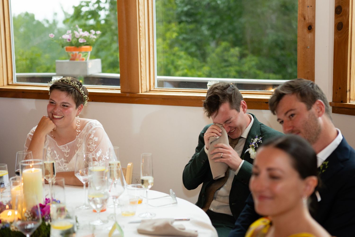 Wedding guests share an emotional moment during dinner reception at a venue with large windows.