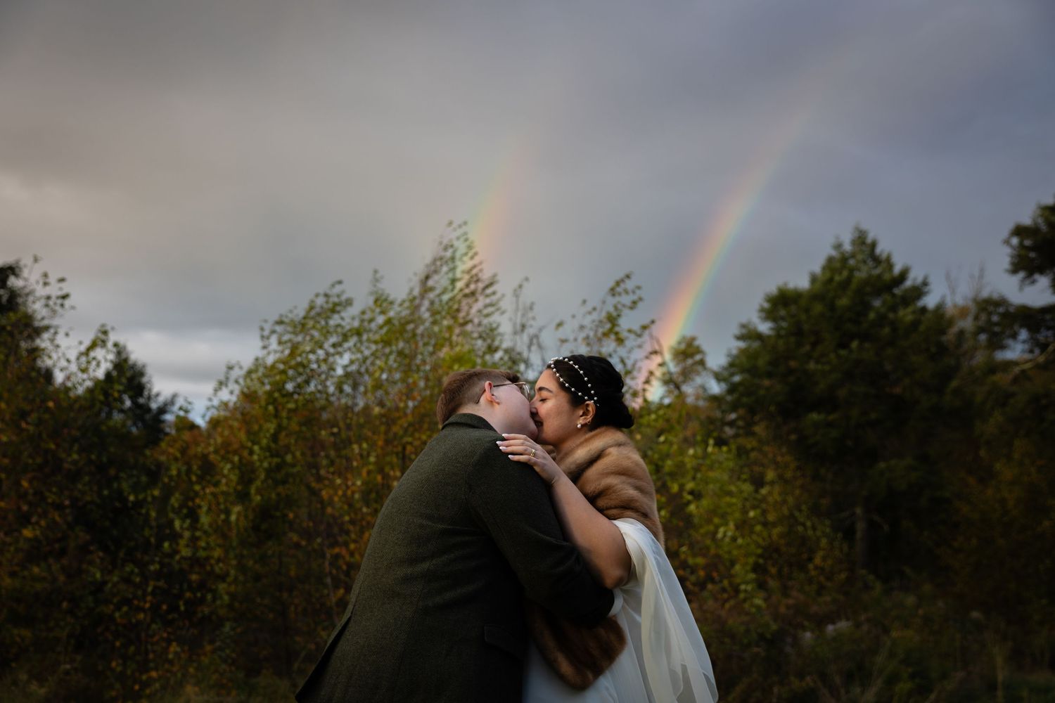 A couple shares an intimate moment under a double rainbow in an autumn landscape.