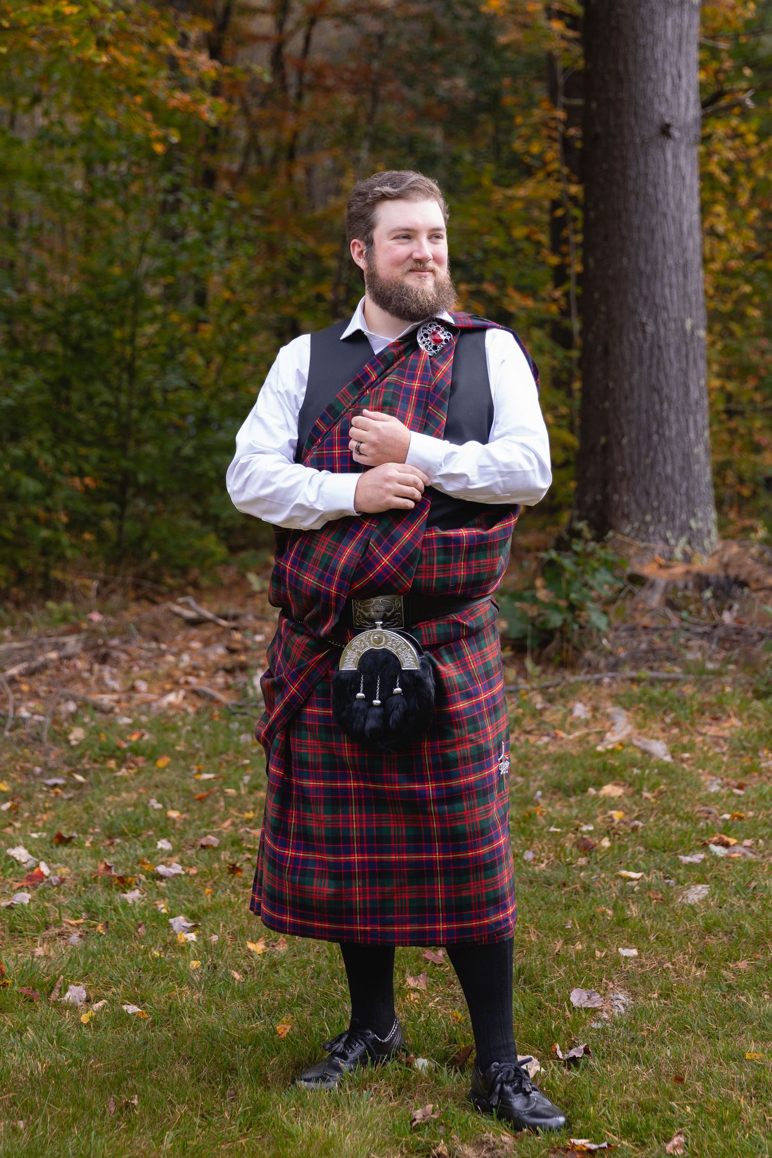 Man wearing traditional Scottish kilt and sporran poses in autumn forest setting.