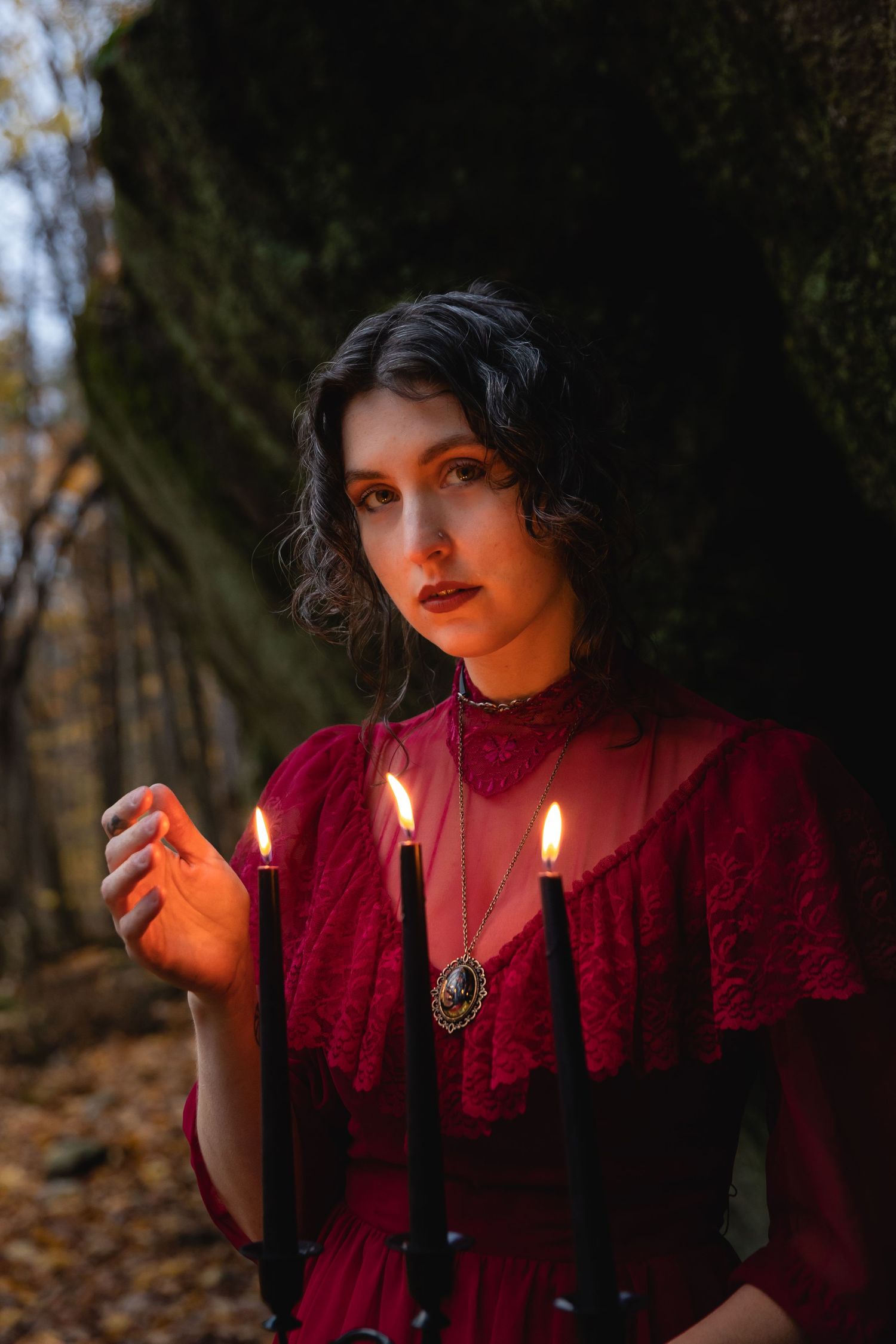Dark-haired woman in red Victorian dress holding black candles in autumn forest.