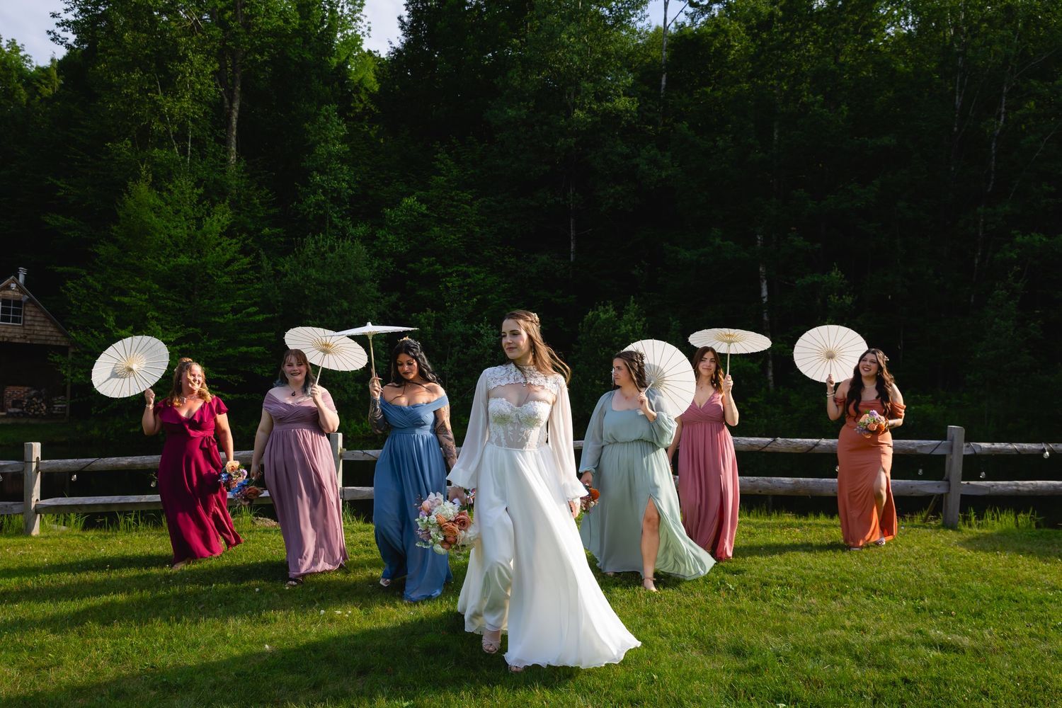 Bride and colorful bridesmaids holding white parasols walking across a green lawn at sunset.