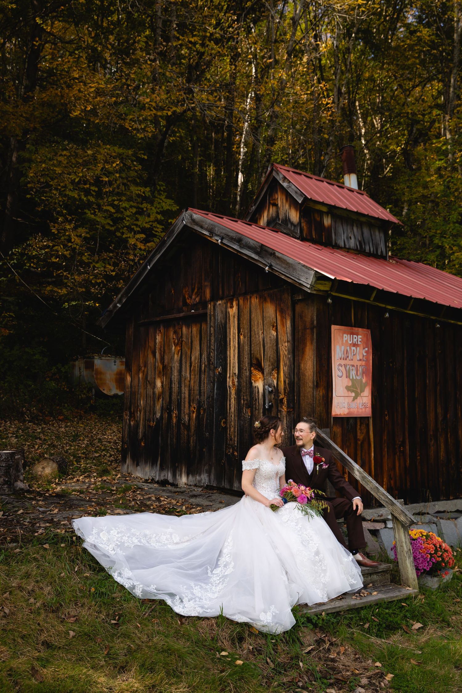 Bride and groom sitting together outside a rustic wooden barn surrounded by colorful fall forest.
