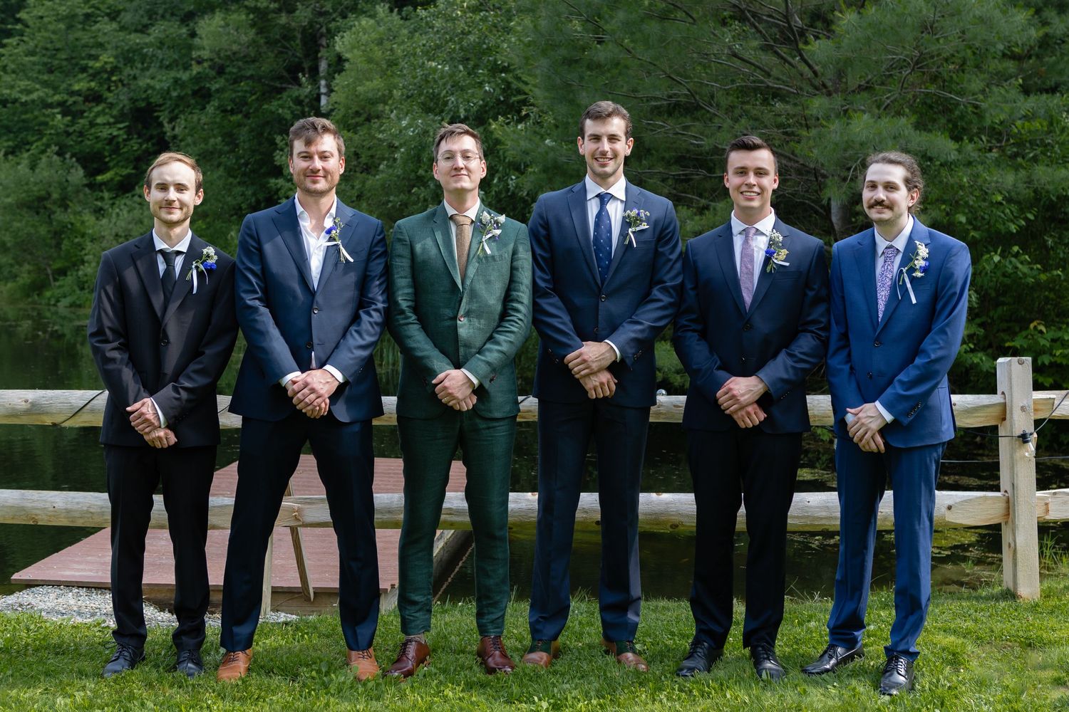 Groomsmen in varied colored suits pose together in front of wooden fence and forest.