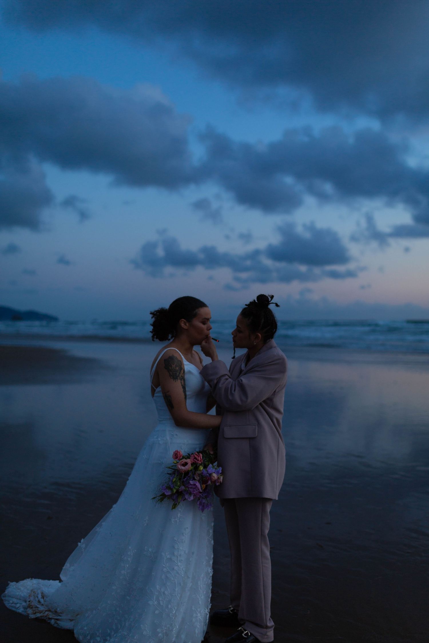 A couple shares an intimate moment on a misty beach at dusk with dramatic cloudy skies.
