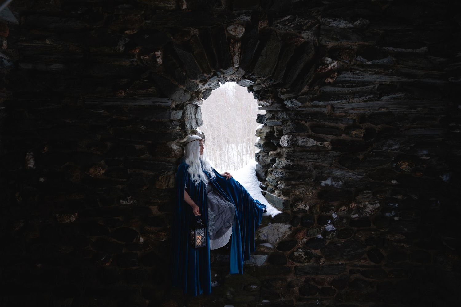 Person in blue cloak standing in stone archway opening to snowy landscape.