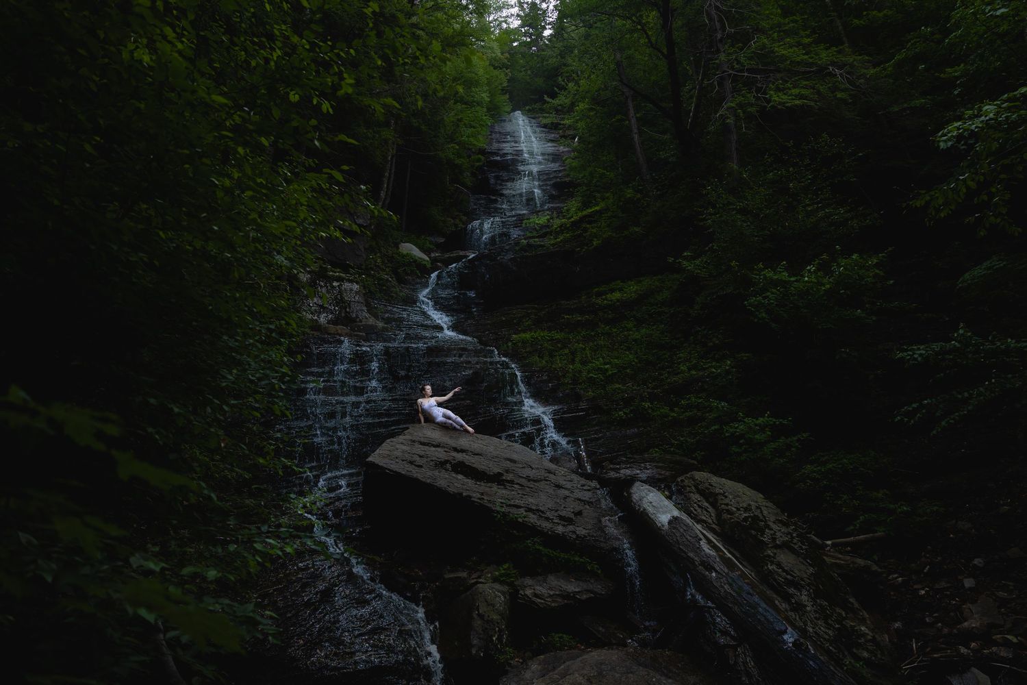 A dancer poses gracefully on rocks beside a cascading waterfall surrounded by lush green forest.