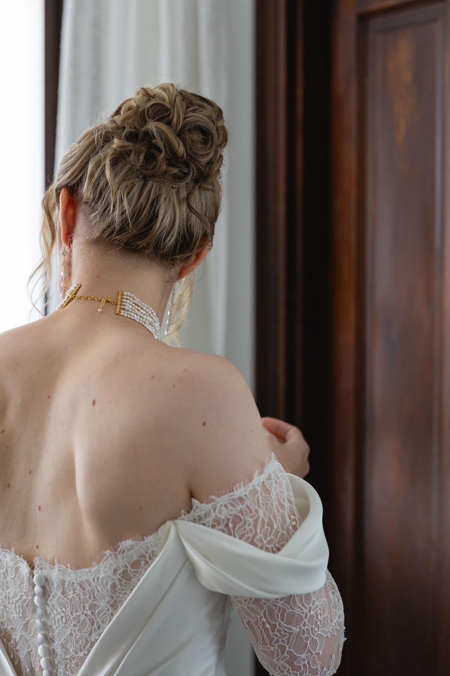 Back view of an elegant bridal look with lace wedding dress and upswept hairstyle against a curtained window.