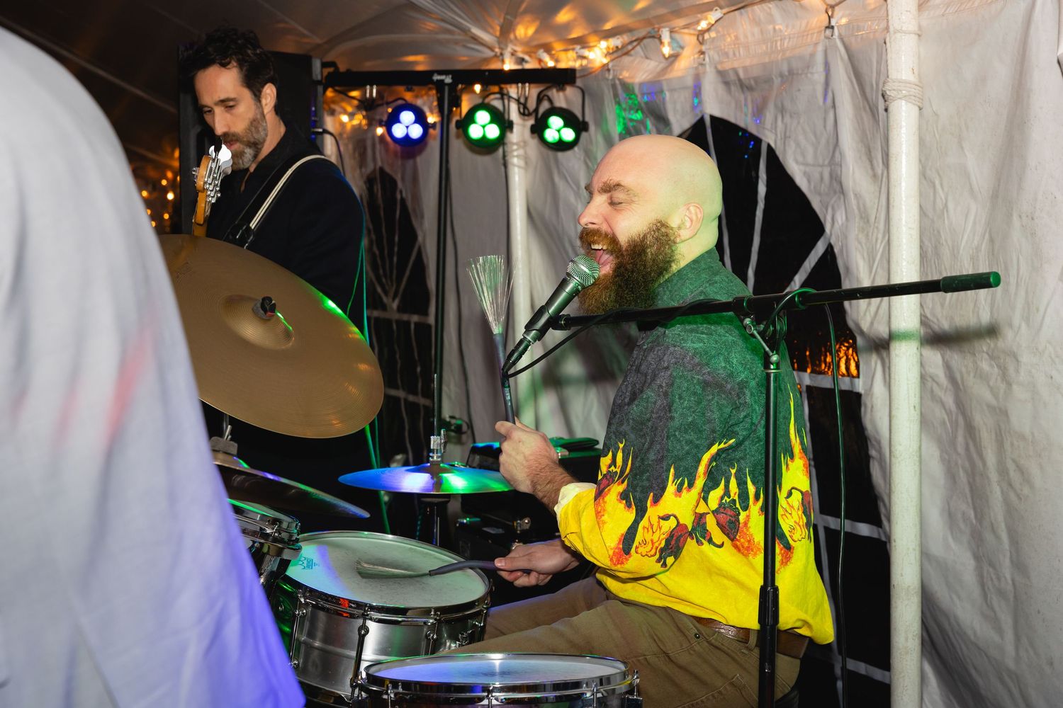 A drummer performs on stage with colorful lighting while other musicians play instruments in the background.