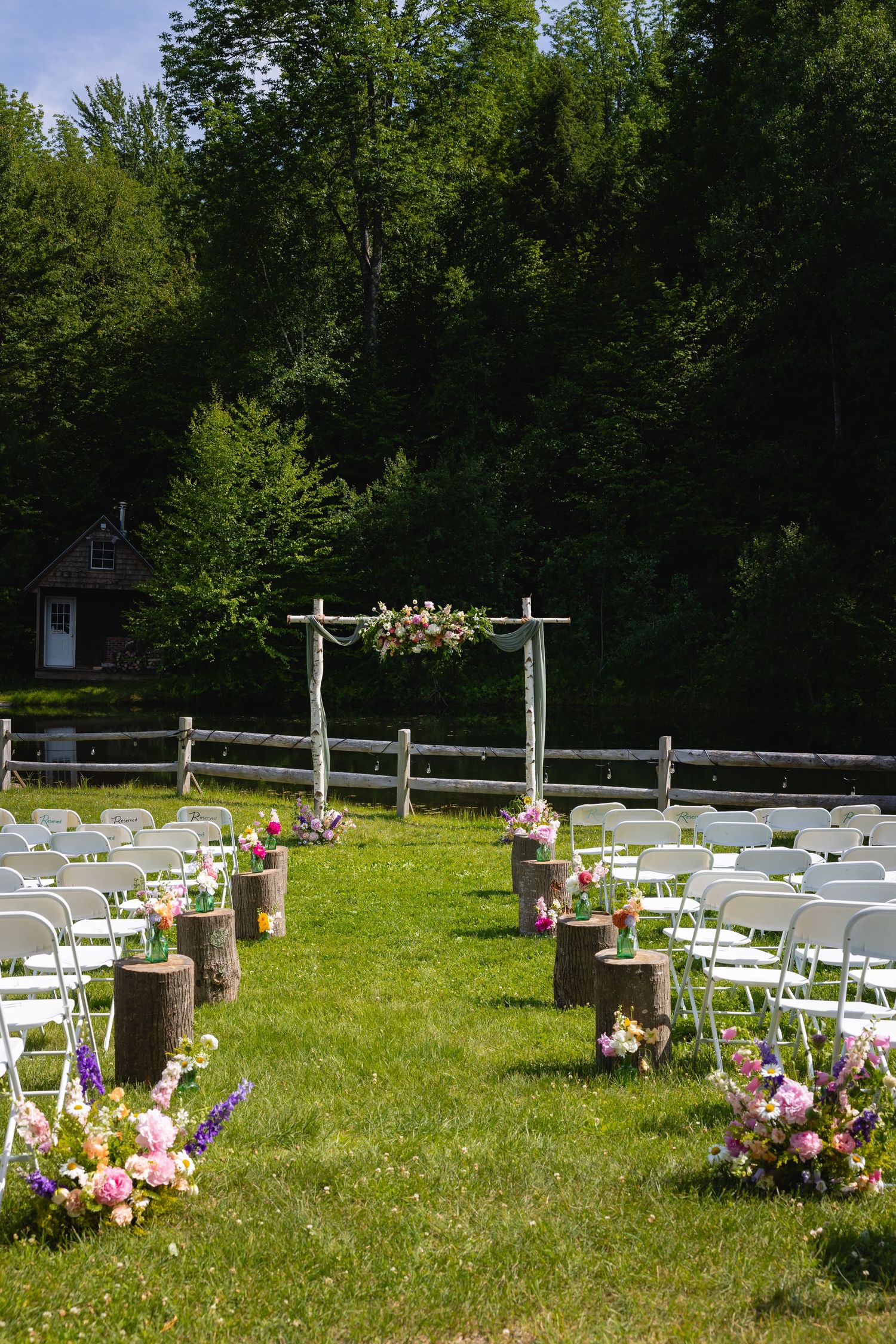 White folding chairs arranged on a grassy lawn for an outdoor wedding ceremony with pink floral decorations and wooden posts.