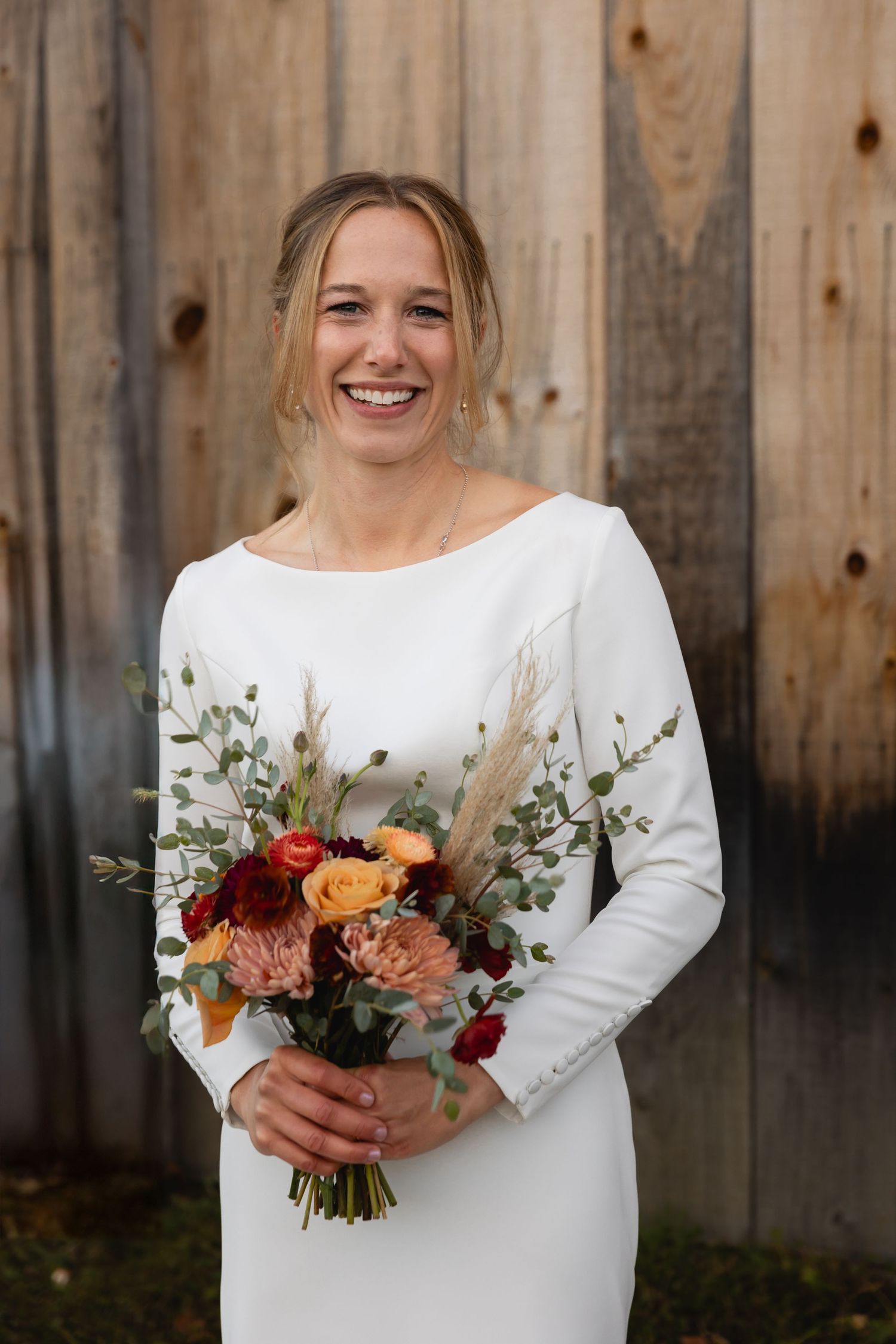 Someone in a simple long-sleeved white dress holds a beautiful autumn bouquet with peach and burgundy flowers against a wood wall.