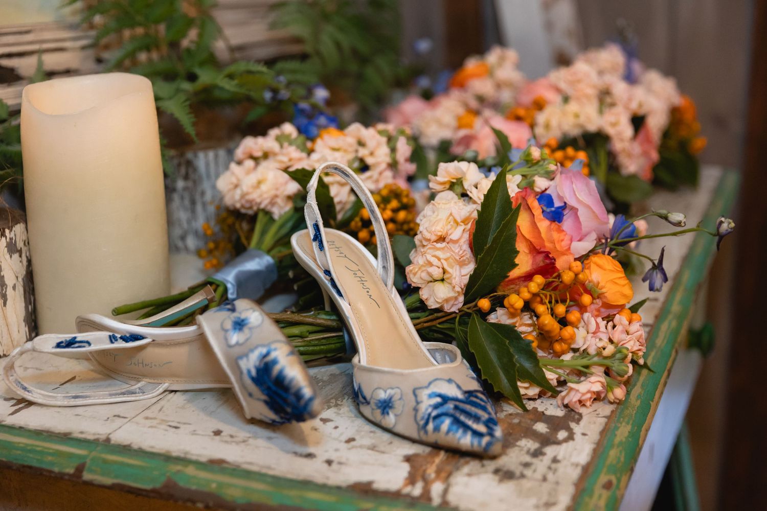 Elegant bridal shoes and floral bouquet with peach and blue flowers arranged on rustic wooden table with white candle.