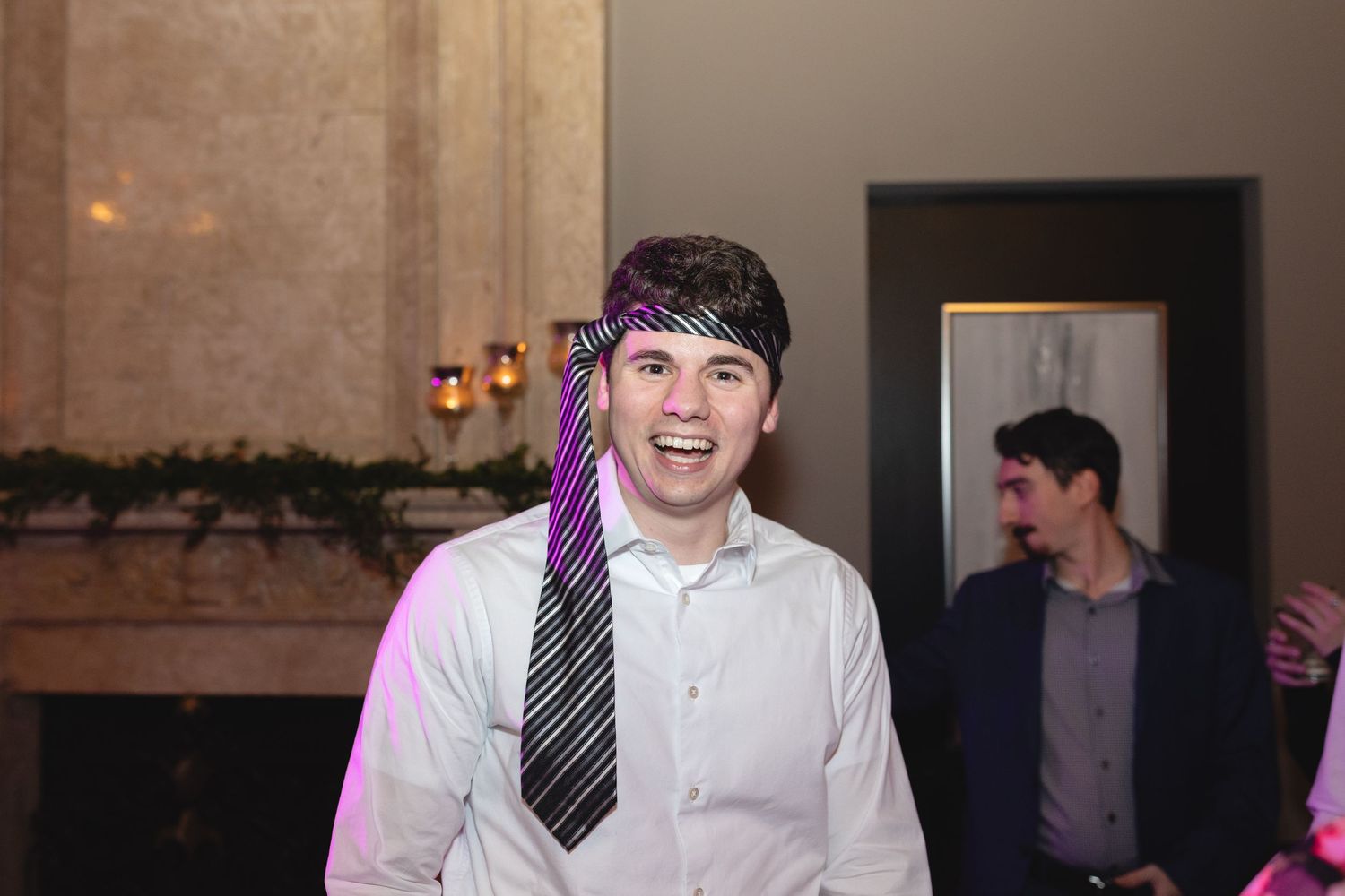 A party guest in a white dress shirt and black tie smiles brightly under purple lighting at an indoor event.
