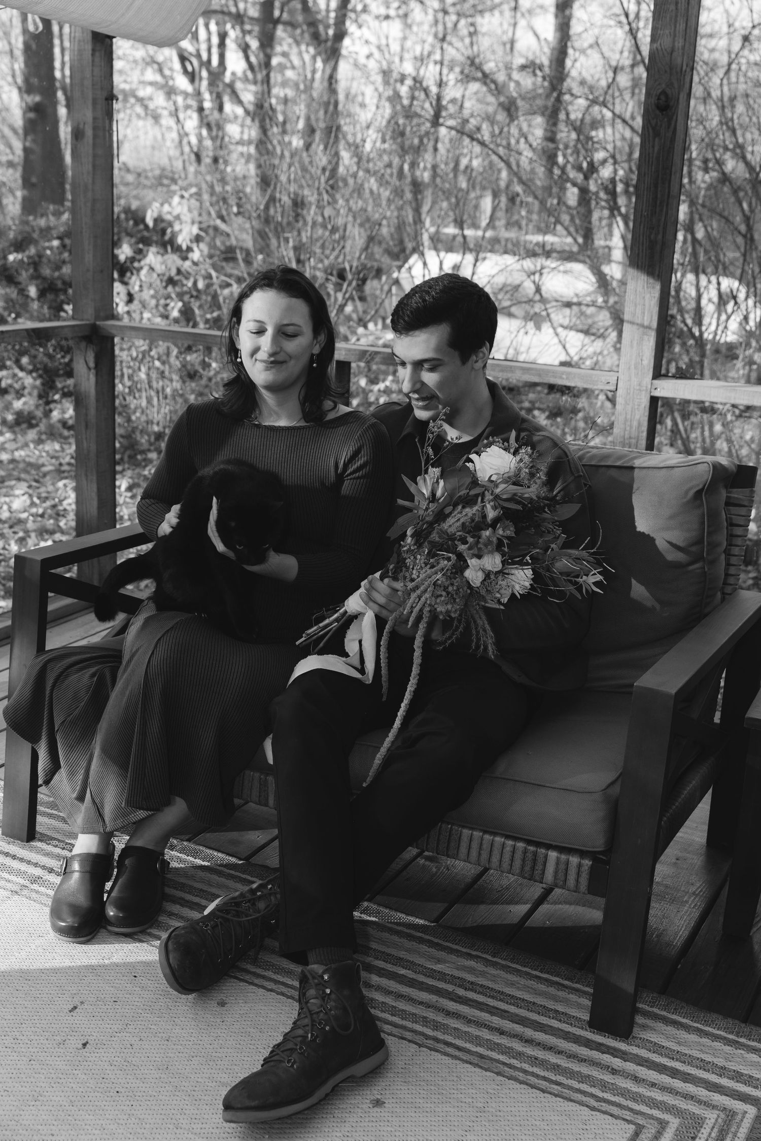 A vintage black and white photograph of a couple sitting together on a porch bench, with one holding a bouquet of flowers.