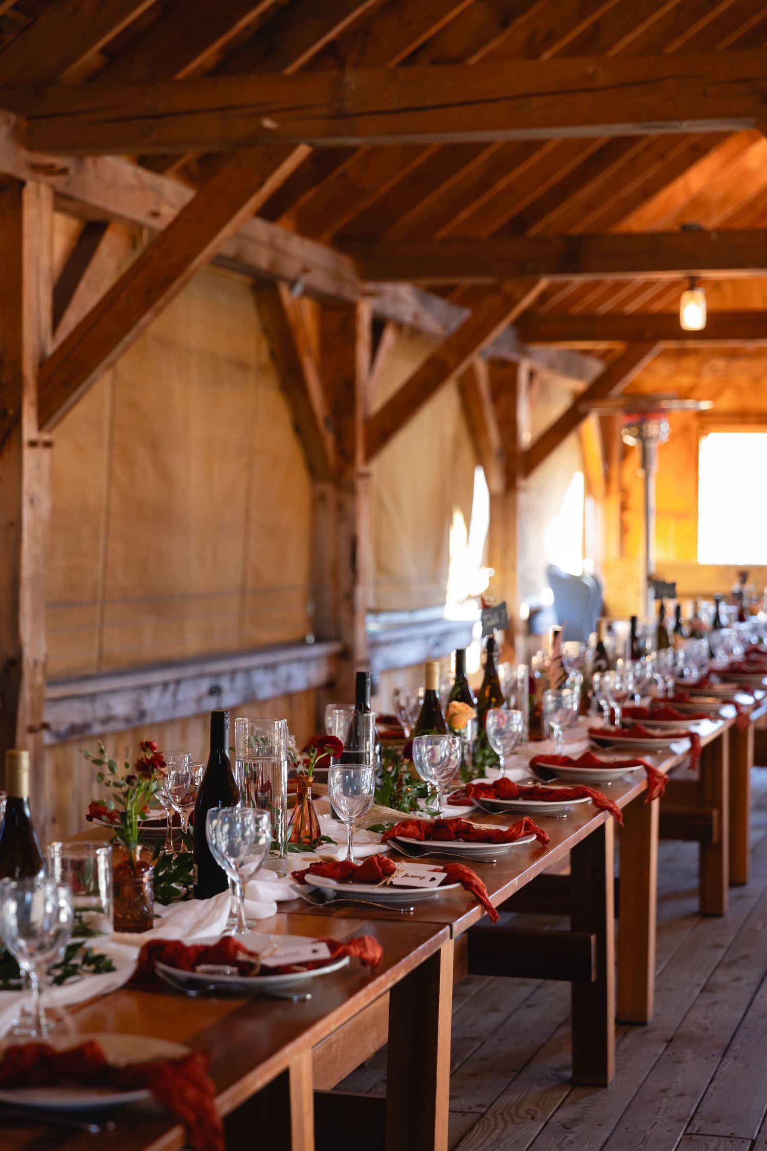 Rustic farm table set for elegant dining with red napkins and wine glasses in a wooden barn with natural lighting.