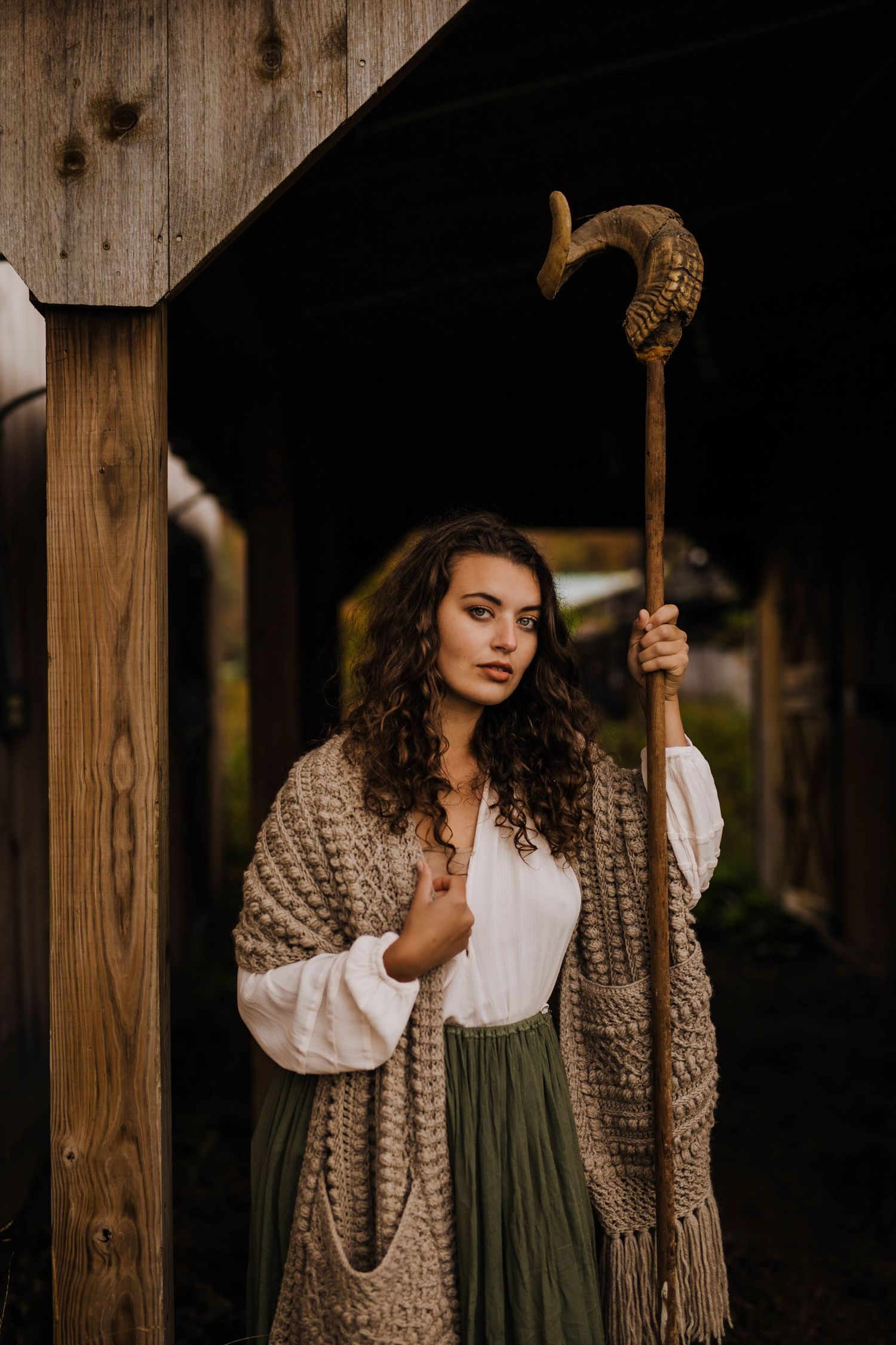 Woman in bohemian outfit holding ornate staff with ram horn topper beside rustic wooden structure.
