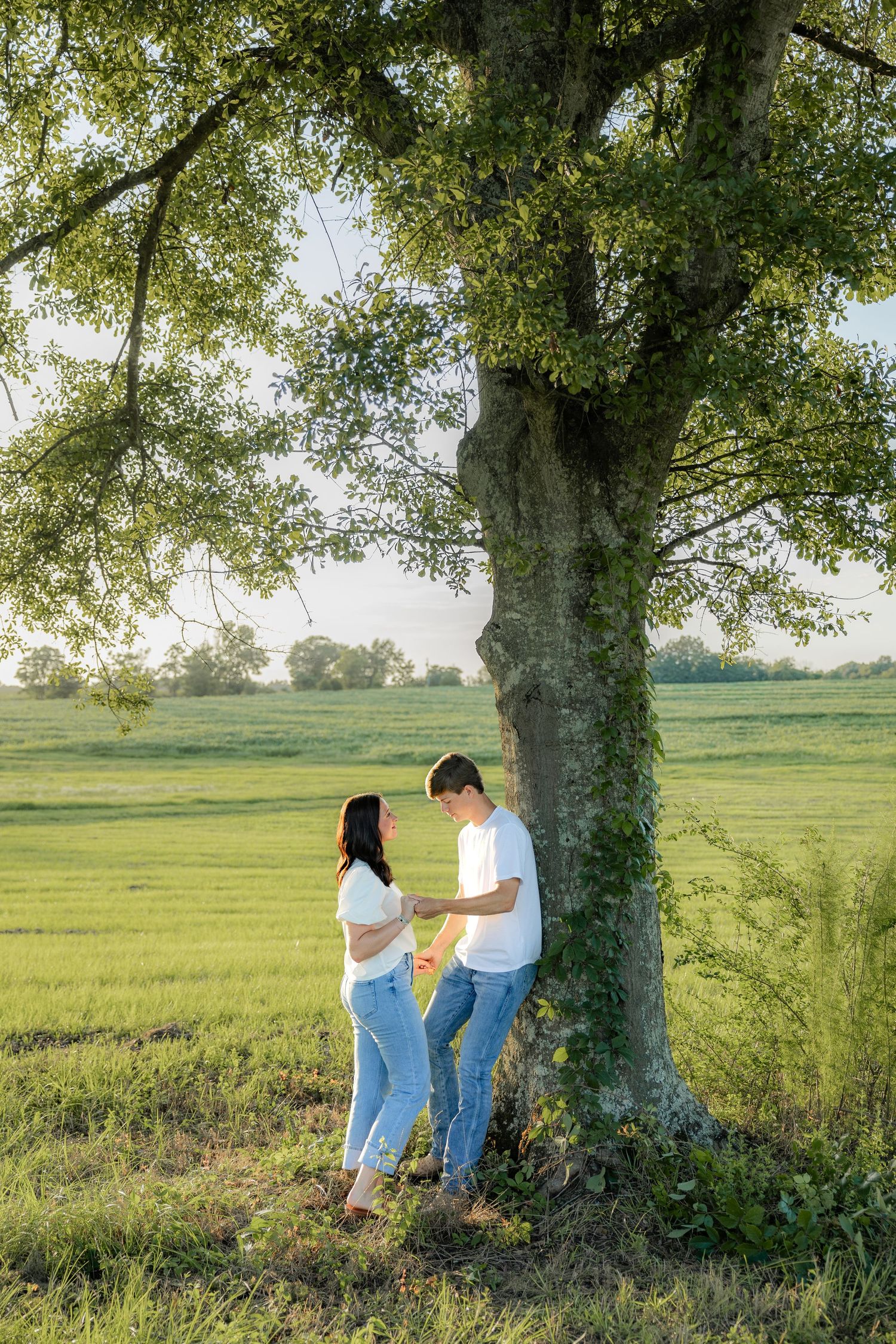 Romantic couple photos in a natural grass field setting with large trees at sunset.