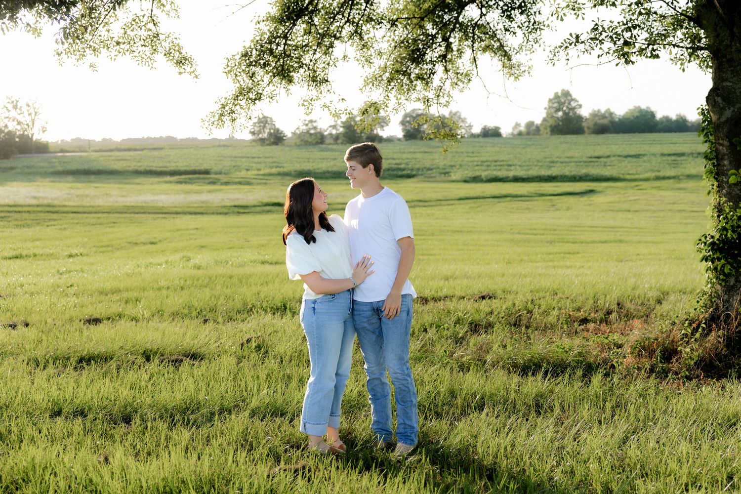 Romantic couple photos in a natural grass field setting with large trees at sunset.