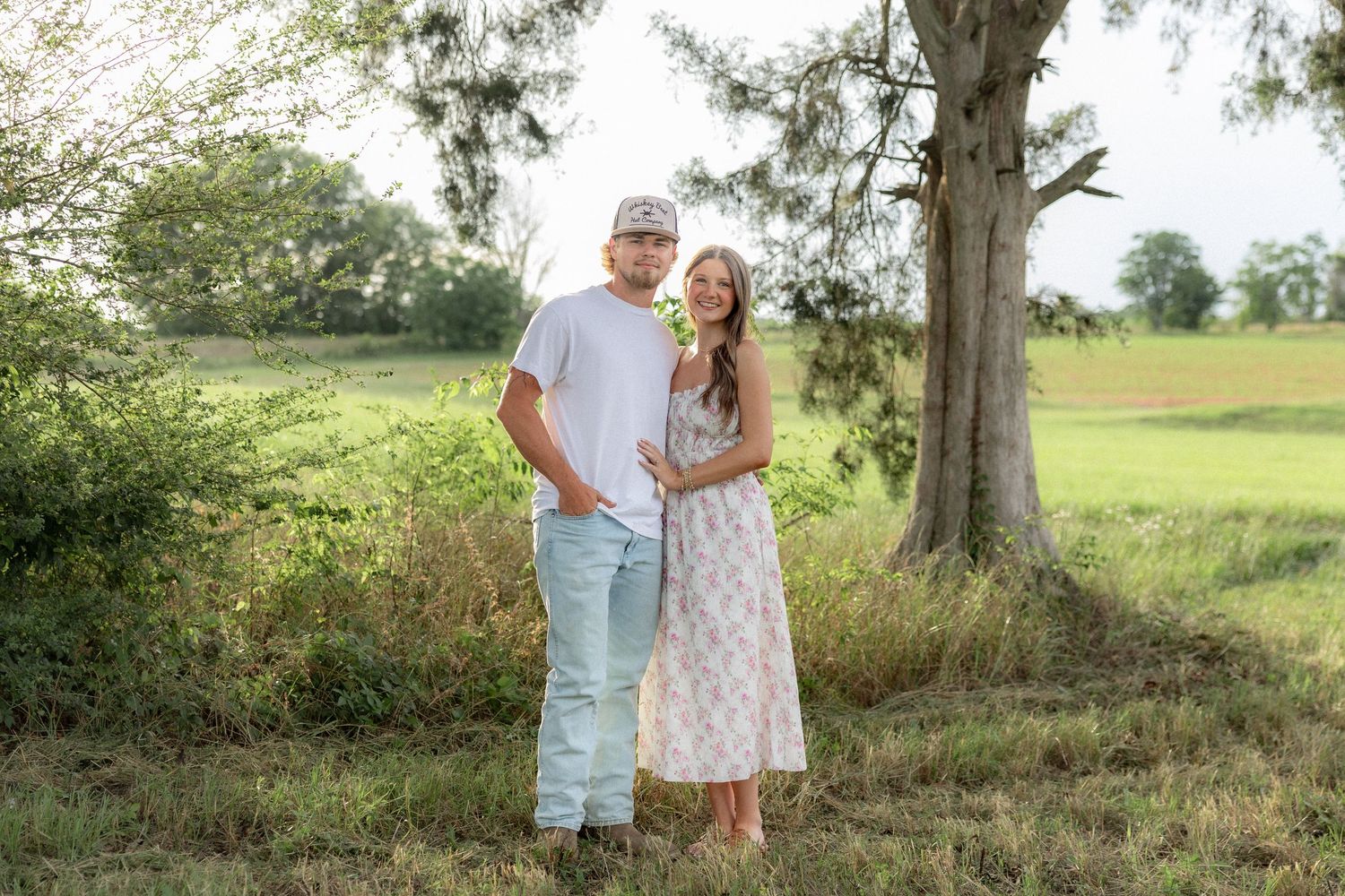 Romantic couple photos in a natural grass field setting with large trees at sunset.