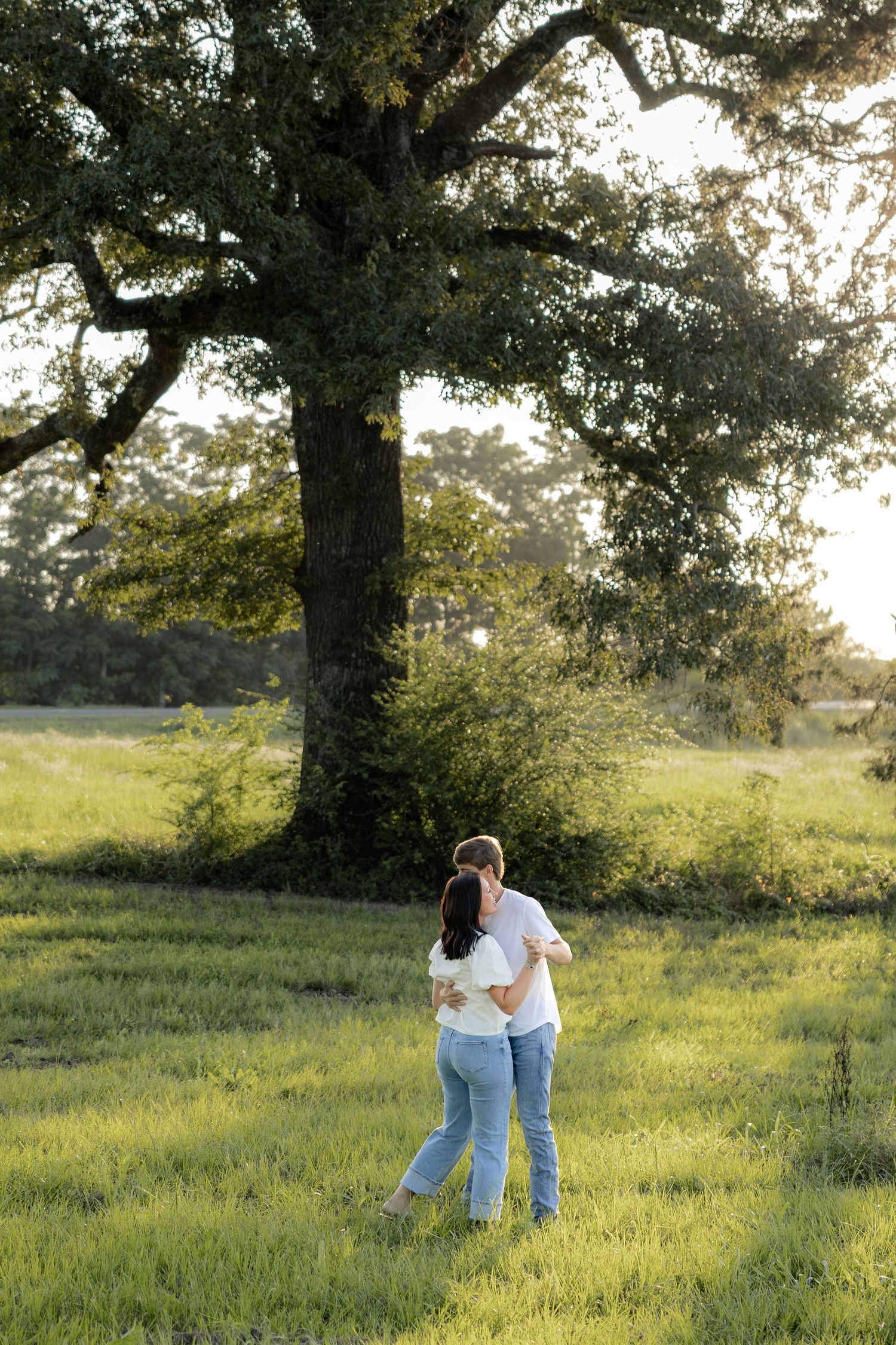 Romantic couple photos in a natural grass field setting with large trees at sunset.