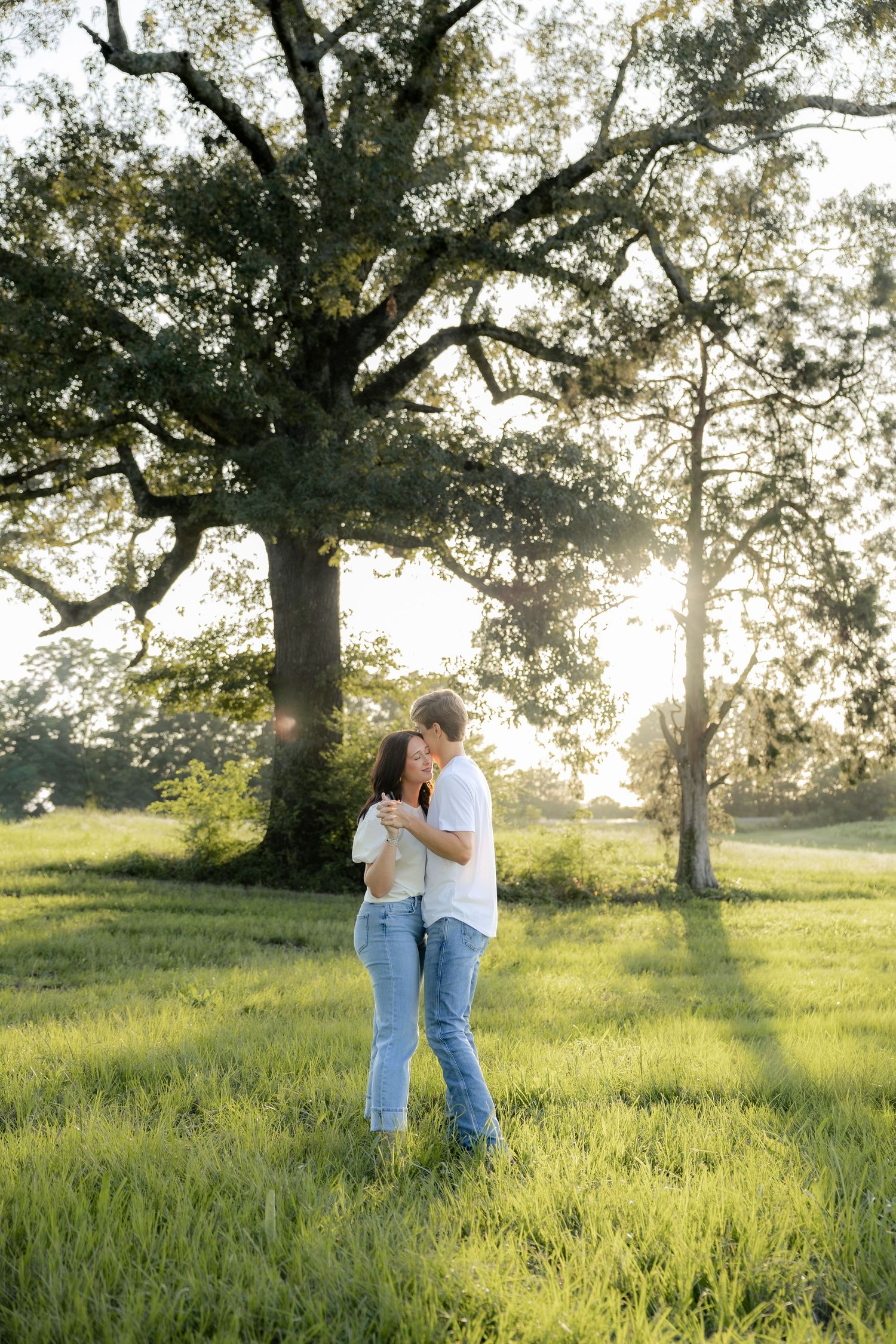 Romantic couple photos in a natural grass field setting with large trees at sunset.