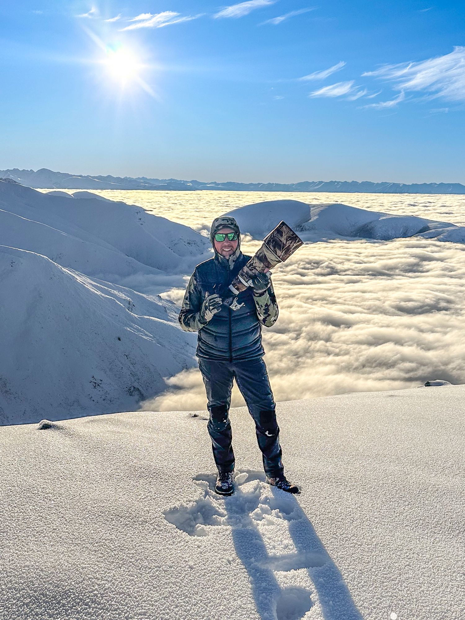 Matt Addington with camera above clouds on a snowy mountain peak under bright sunlight.