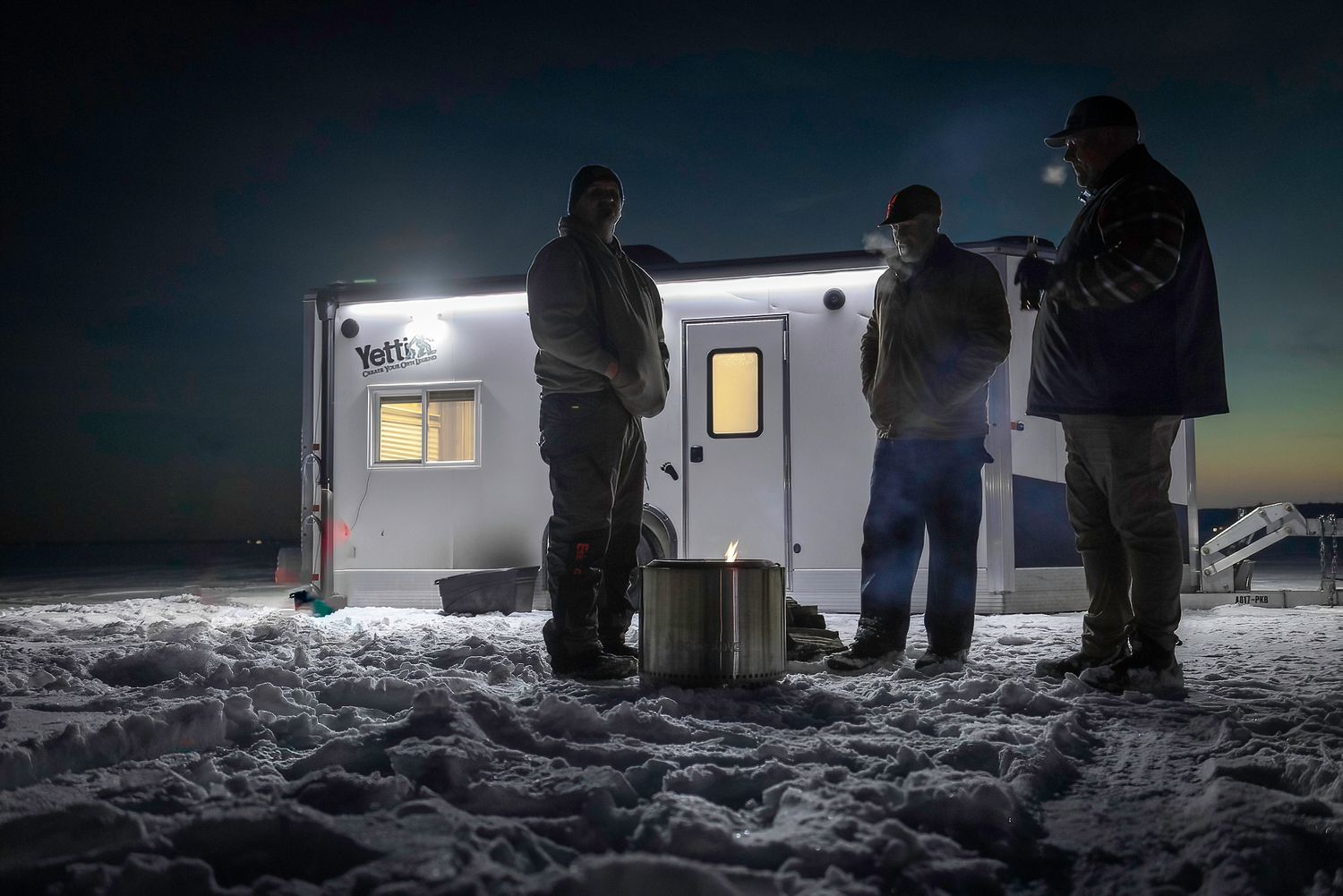 Group gathered outside illuminated ice fishing shelter at night on frozen lake.