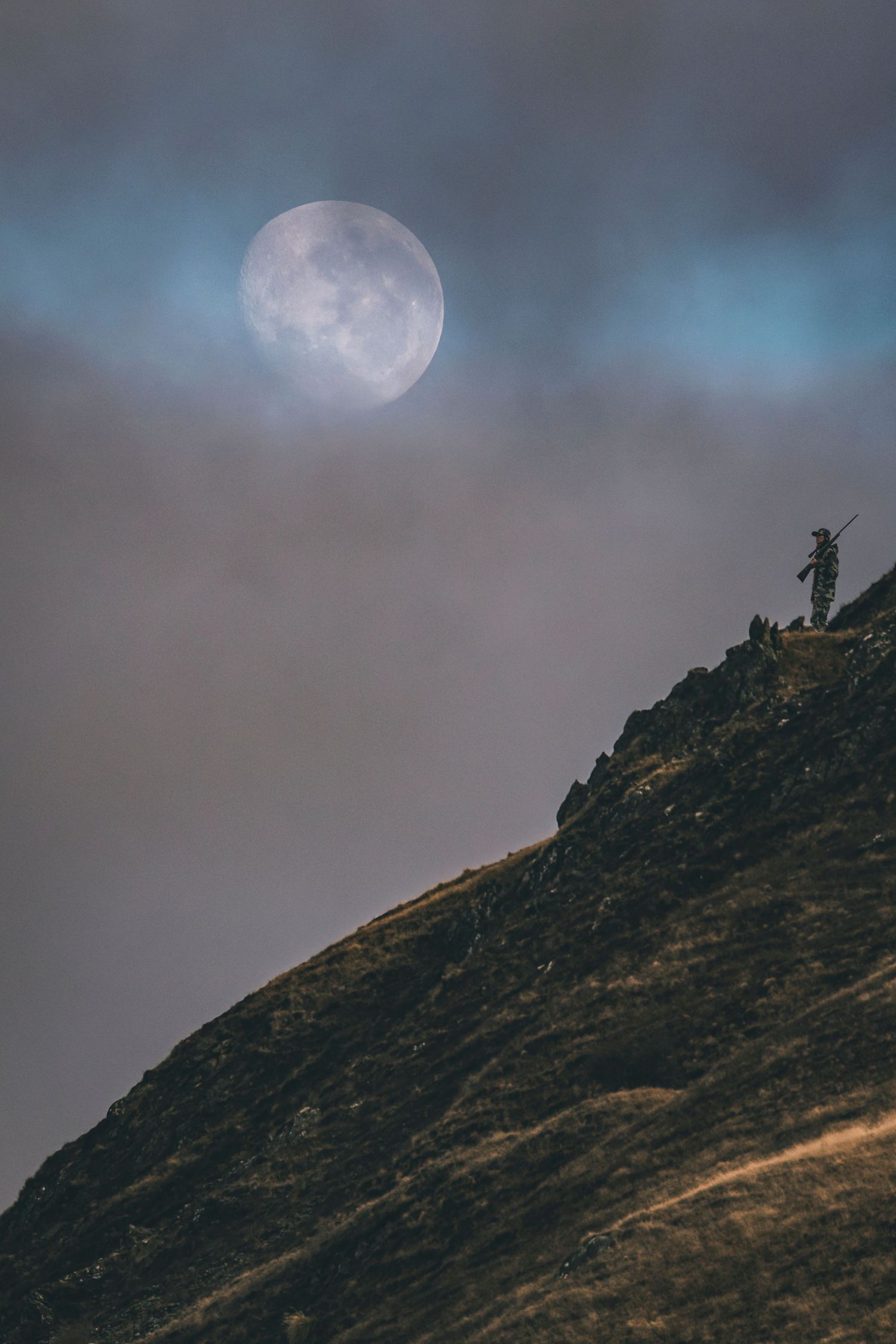 A silhouetted figure stands on a mountain peak beneath a glowing full moon in misty conditions.