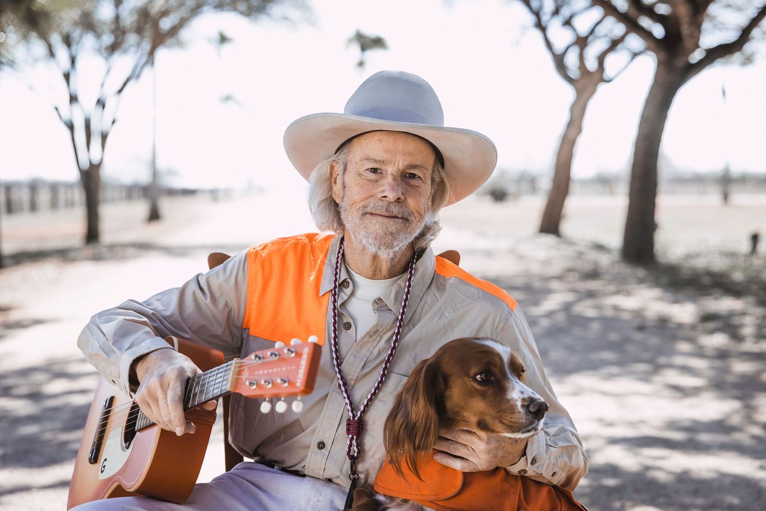 A musician in western attire plays guitar outdoors with a dog by their side.